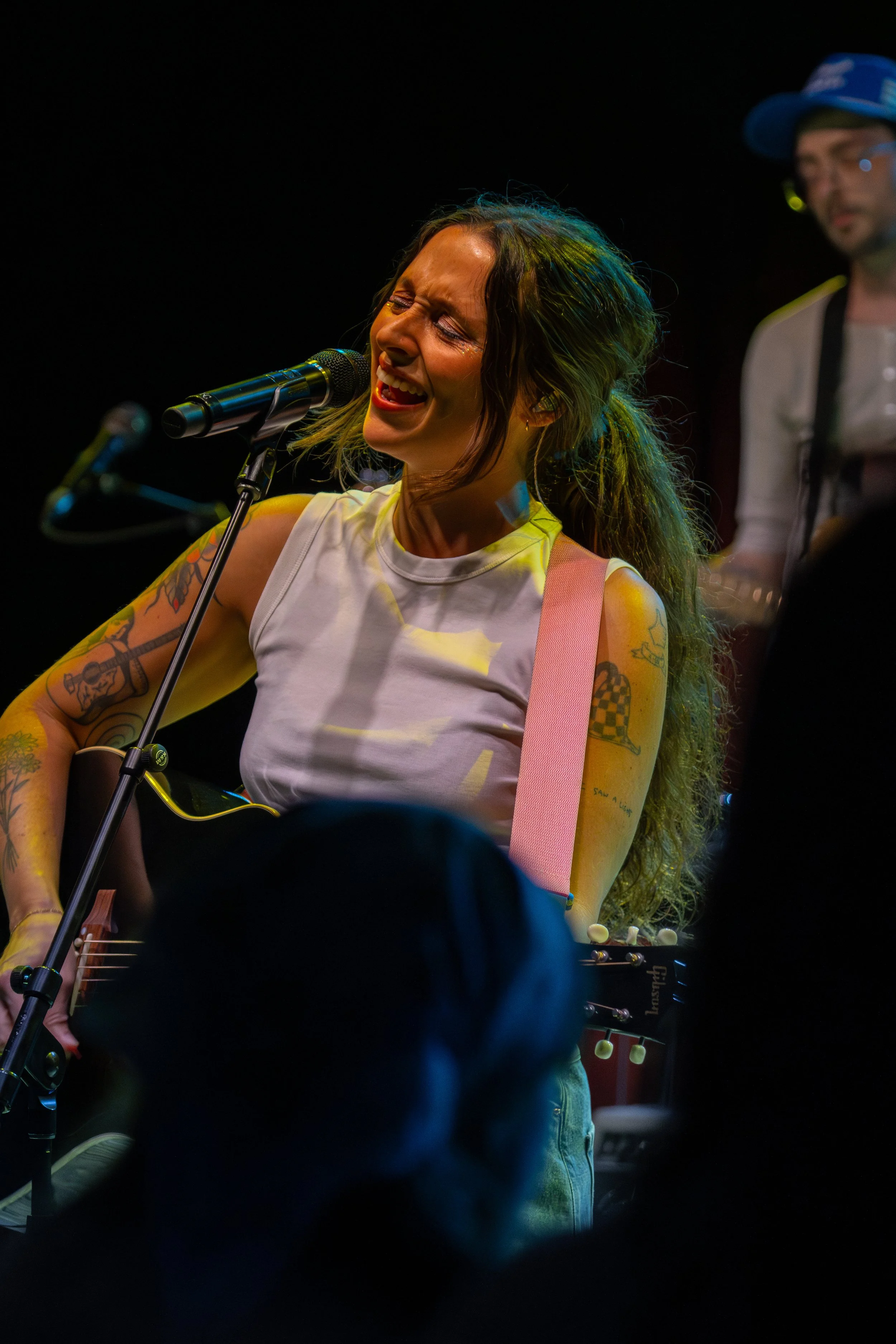 A woman with tattoos singing passionately into a microphone while playing an acoustic guitar, with a man in the background wearing glasses and a blue hat.