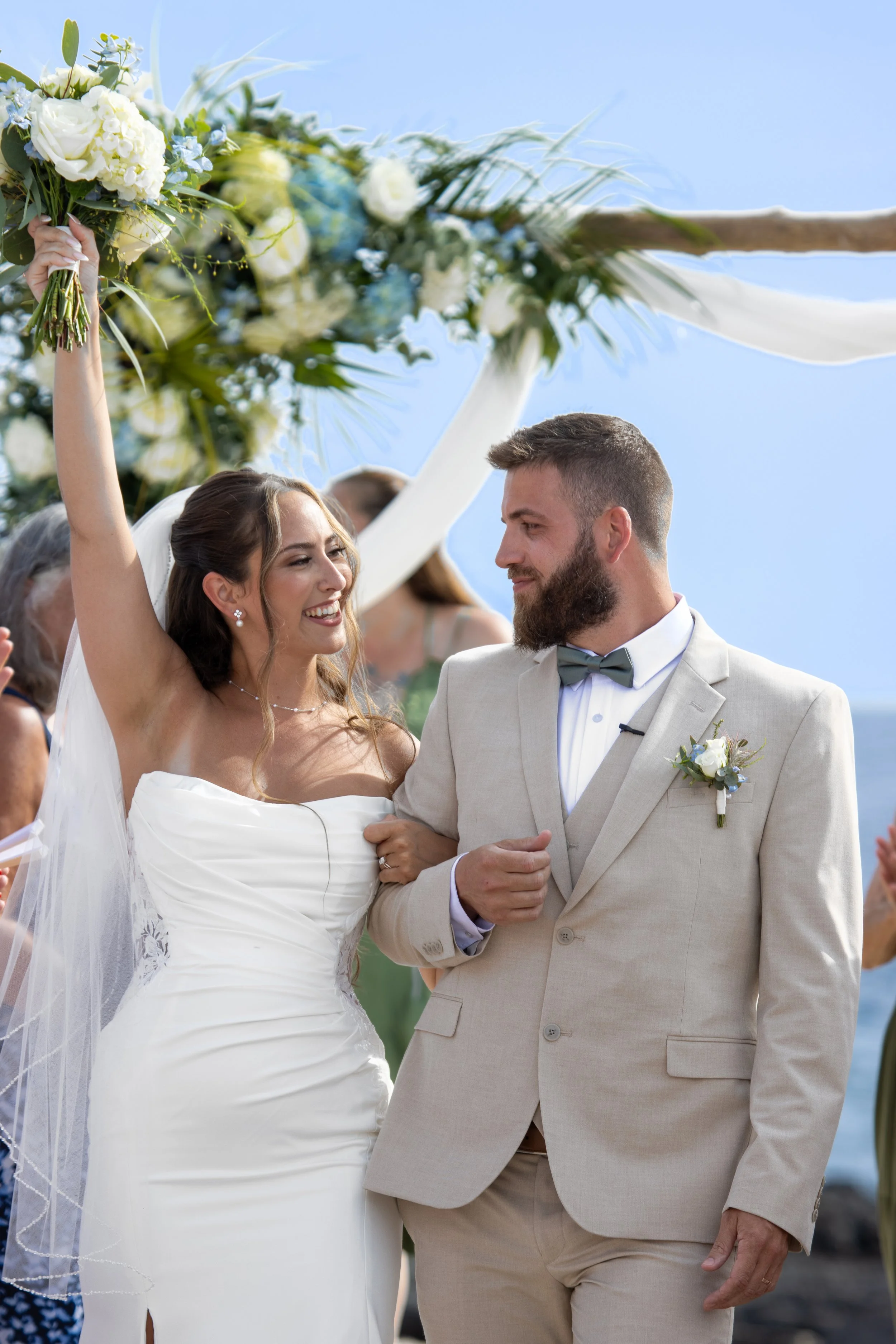 A bride and groom smiling during their wedding ceremony outdoors. The bride is holding a bouquet of white and blue flowers, and the groom is wearing a light-colored suit with a bow tie and boutonniere. They are under a decorated arch with white fabri