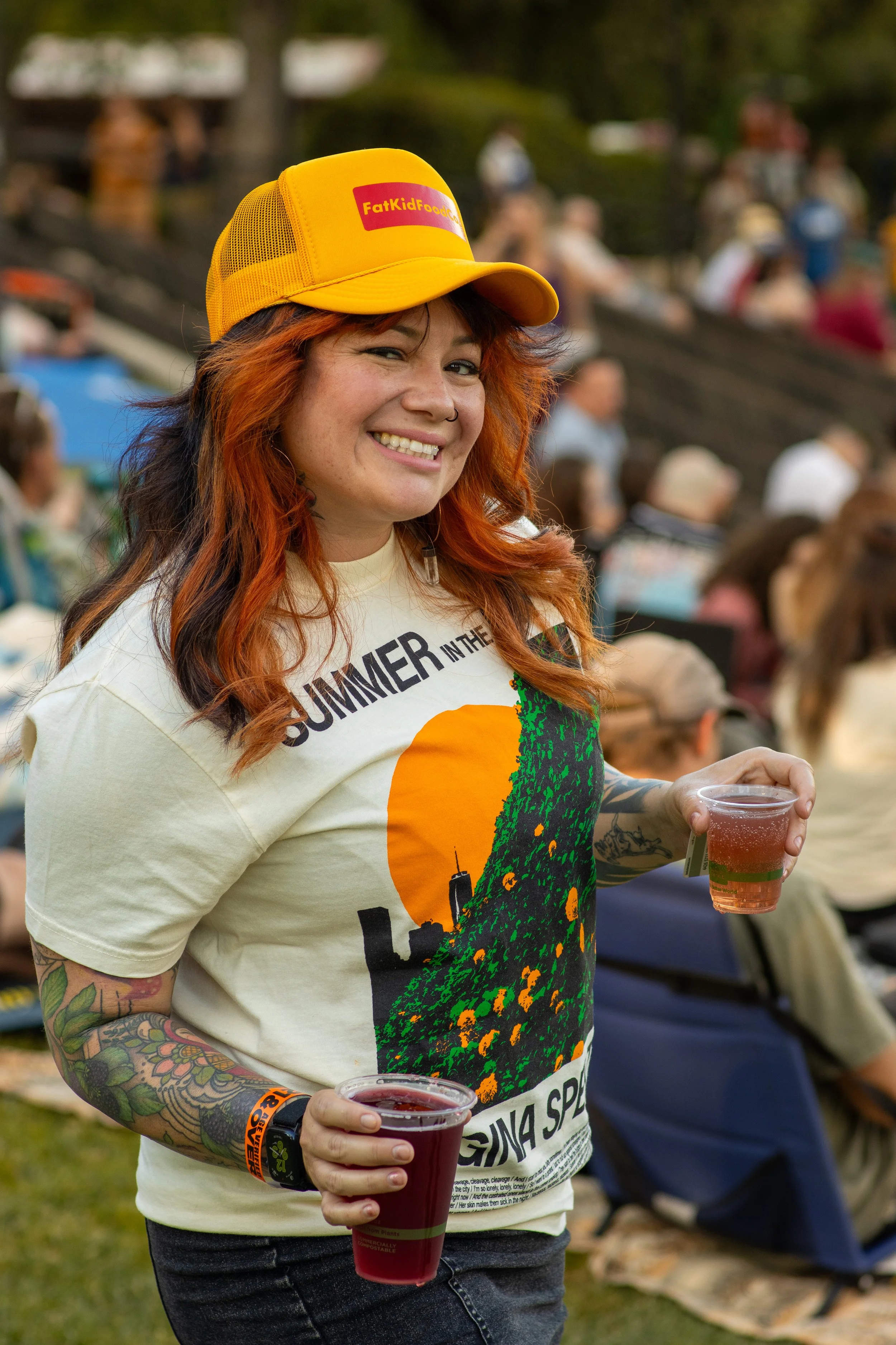 A young woman with red and black hair, tattoos on her arms, and a nose ring, smiling while holding a cup of red drink at an outdoor event. She wears a yellow trucker hat with a red and yellow patch and a T-shirt with colorful graphics. People are blu