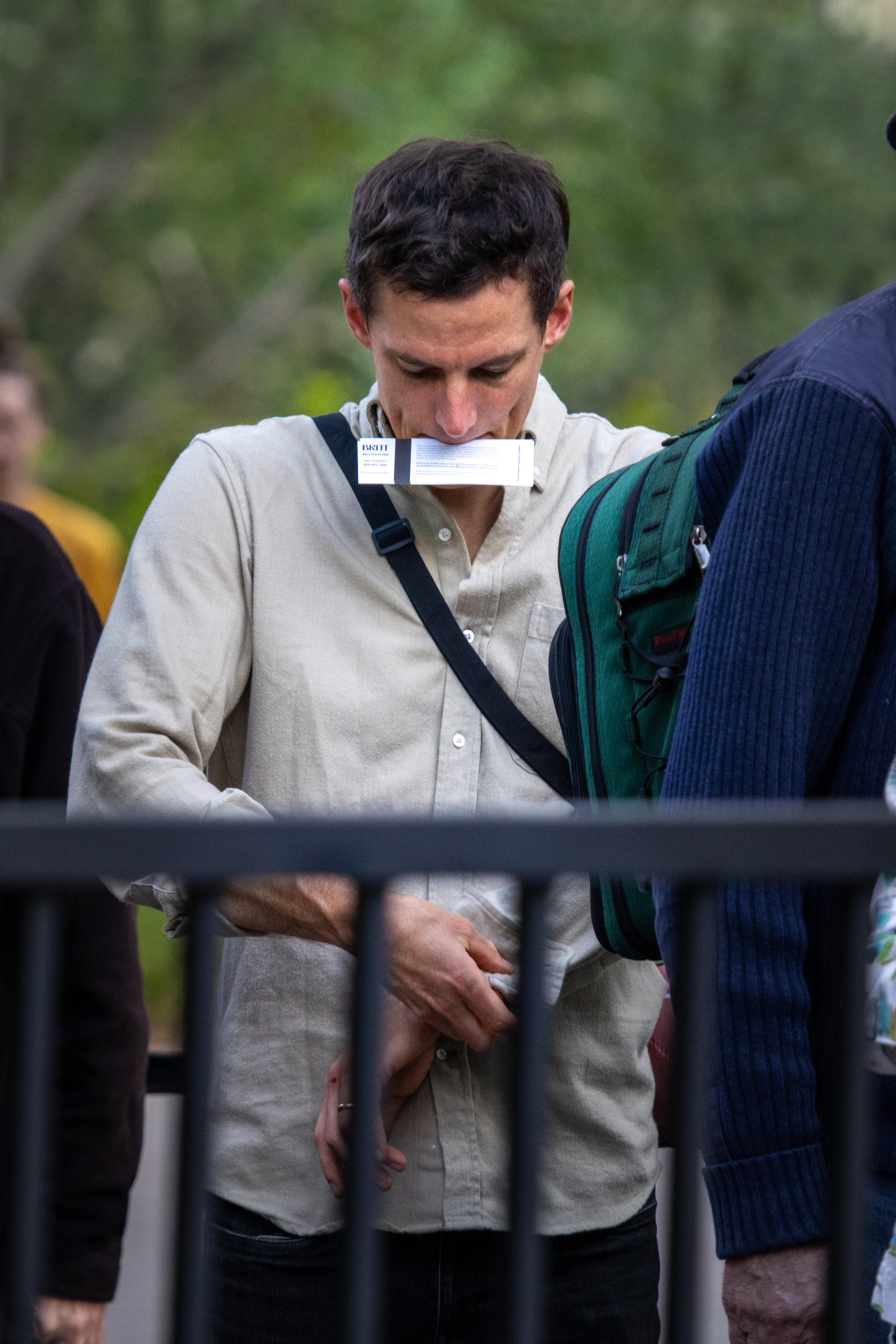 A person standing behind a black iron fence, wearing a beige shirt, holding their hands together in front of them, with their head bowed. There are other people around. The background shows green trees.
