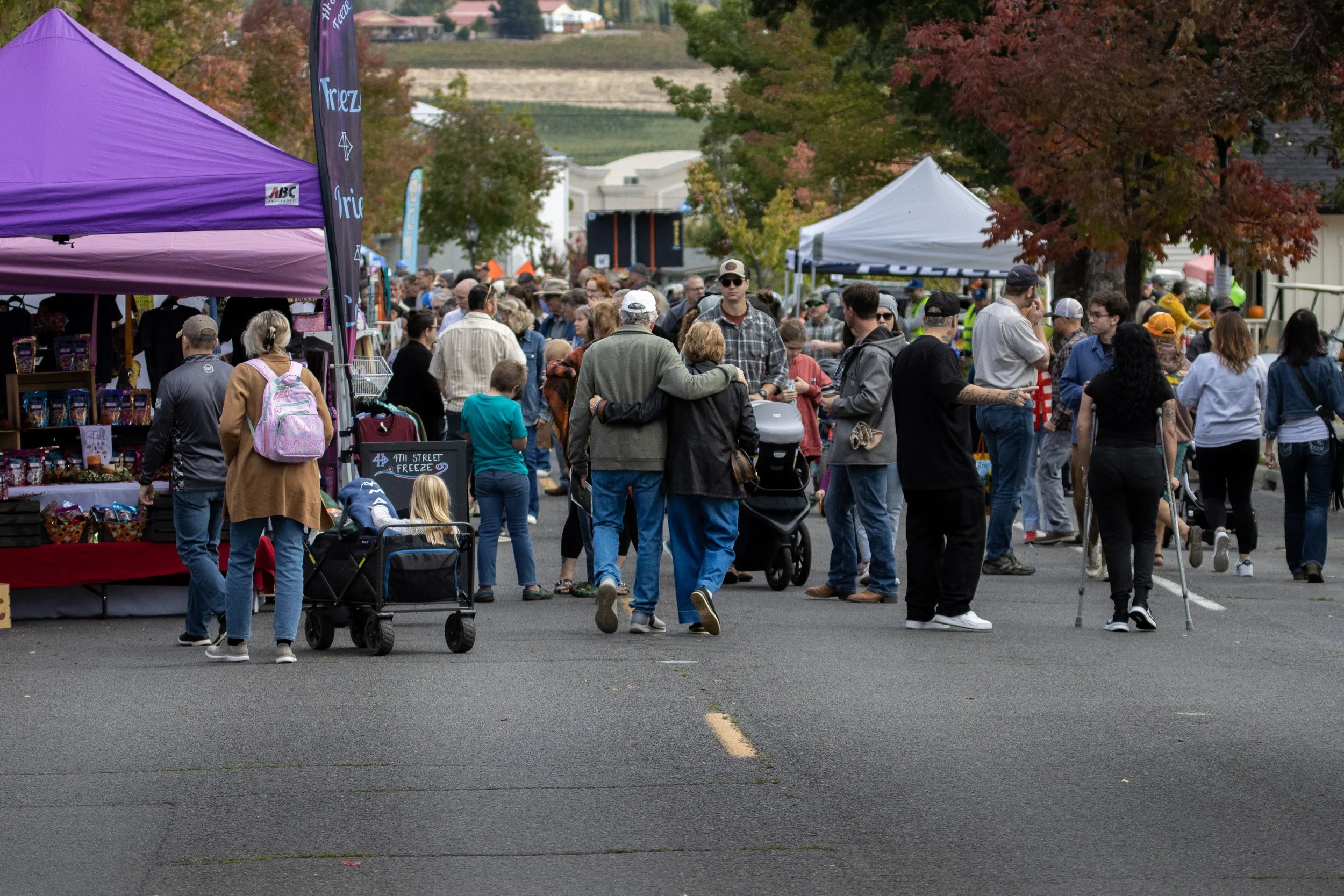 A crowd gathered at an outdoor event with booths, tents, and trees with fall foliage.
