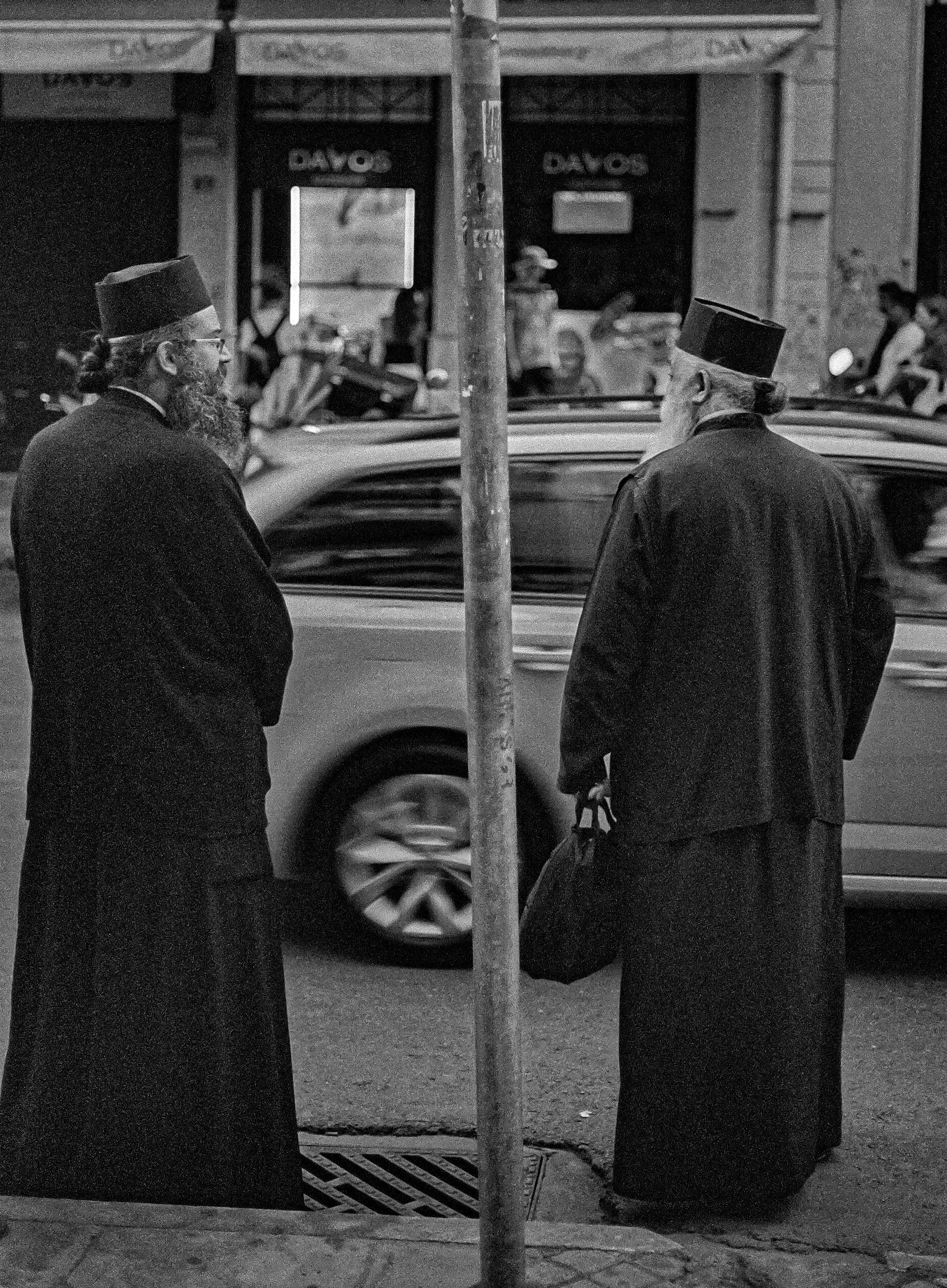 Two men wearing traditional religious clothing and hats are standing on opposite sides of a street, separated by a pole. They are facing each other and appear to be engaged in conversation. Behind them is a car in motion and a blurred background of a