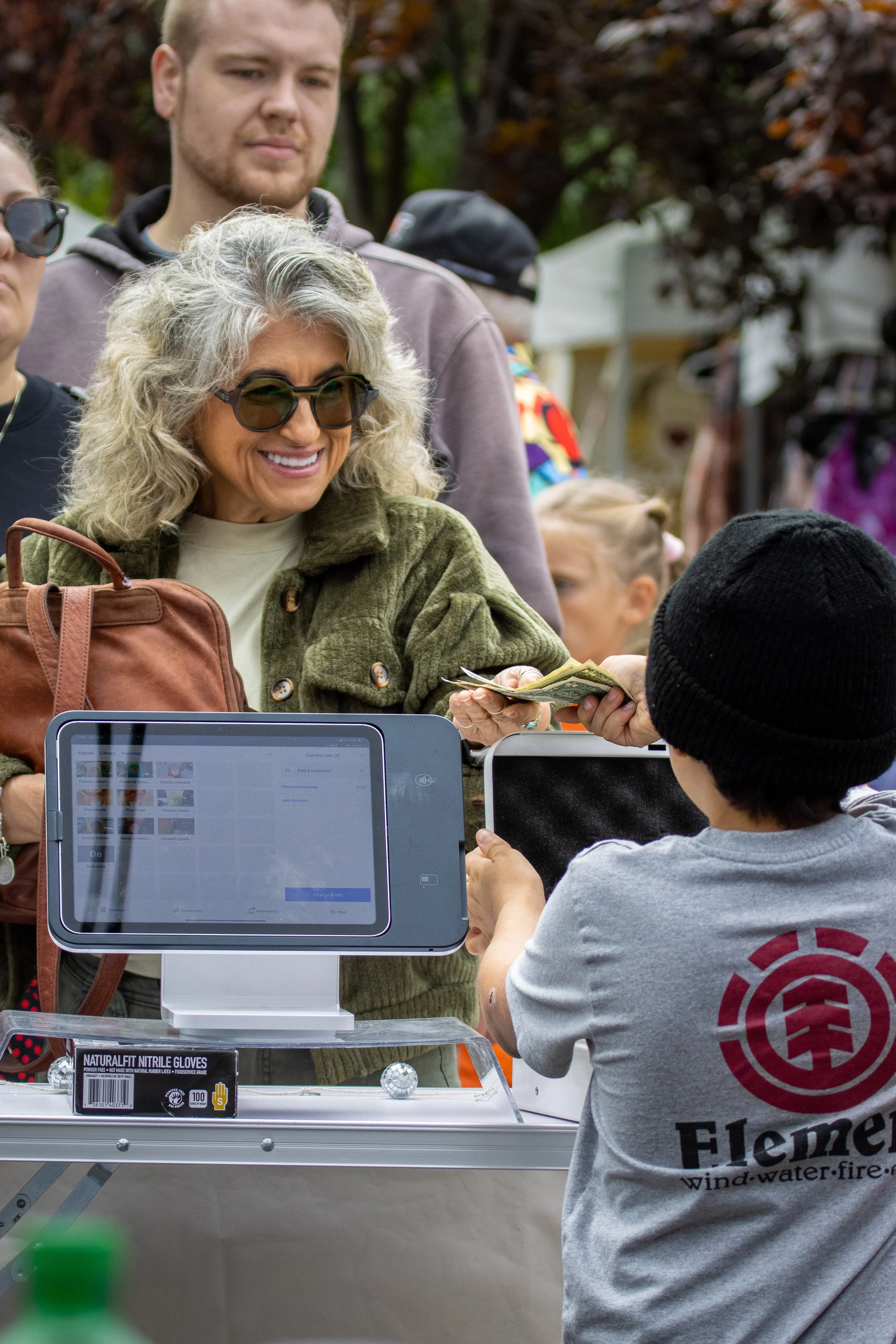 A woman with gray curly hair and sunglasses is smiling as she hands cash to a young man in a gray shirt with a logo, at an outdoor event. The woman is wearing a green jacket and holding a brown purse, while the man is wearing a gray t-shirt with a ci