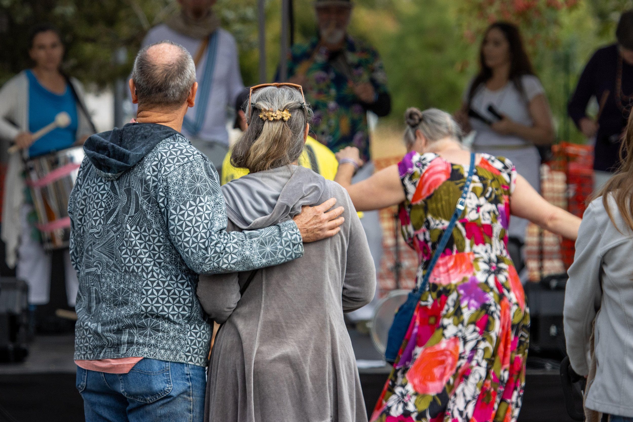 People gathered outdoors in a park, engaged in conversation, with trees and a vendor stand in the background.