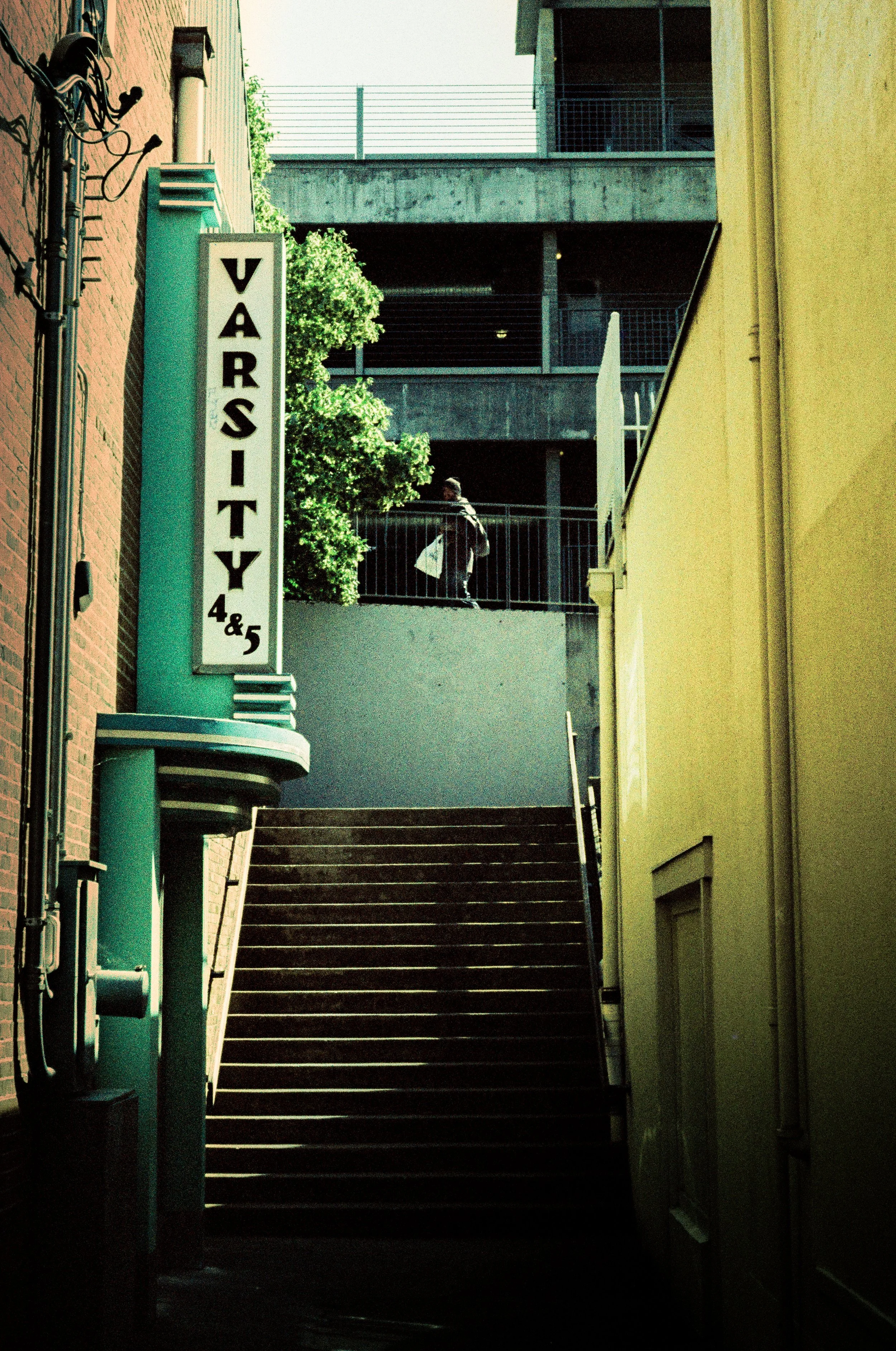 Outdoor staircase leading up to a balcony area at the back of a building, with a person walking and carrying a shopping bag.