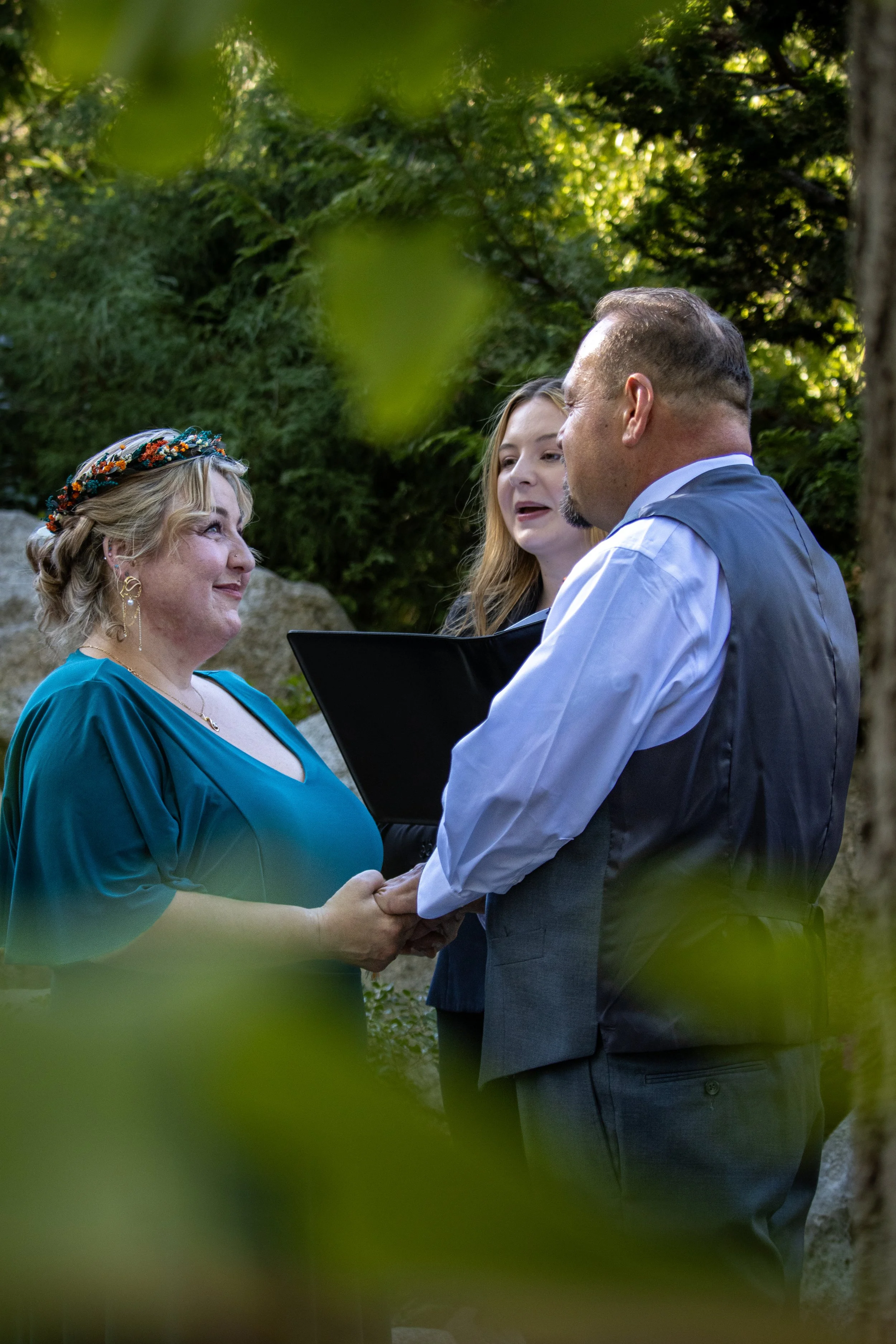A couple getting married outdoors, holding hands and exchanging wedding vows, with an officiant standing beside them, in a lush green garden.