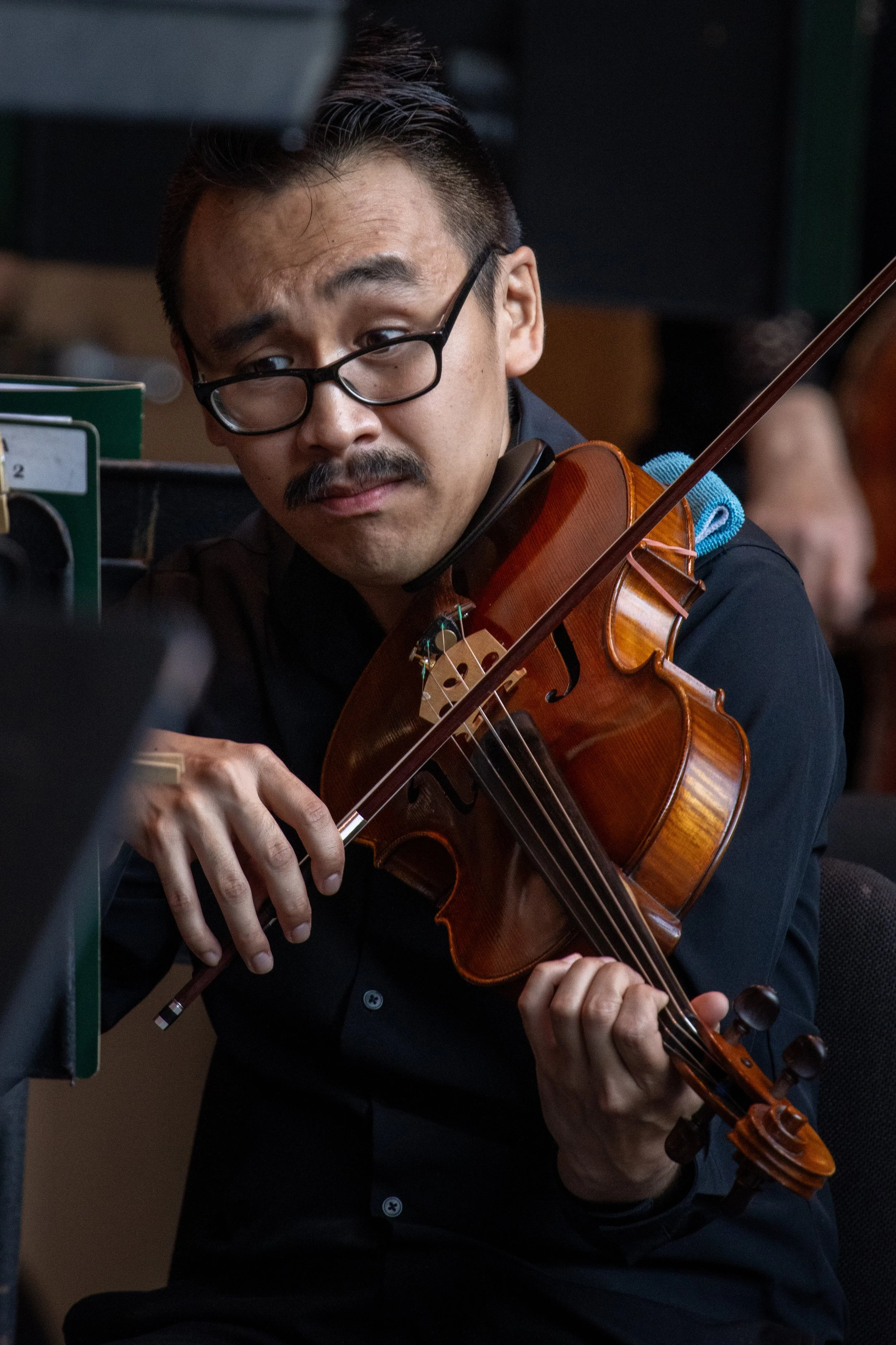 Man wearing glasses playing the violin in a room, with sheet music and a person in the background.