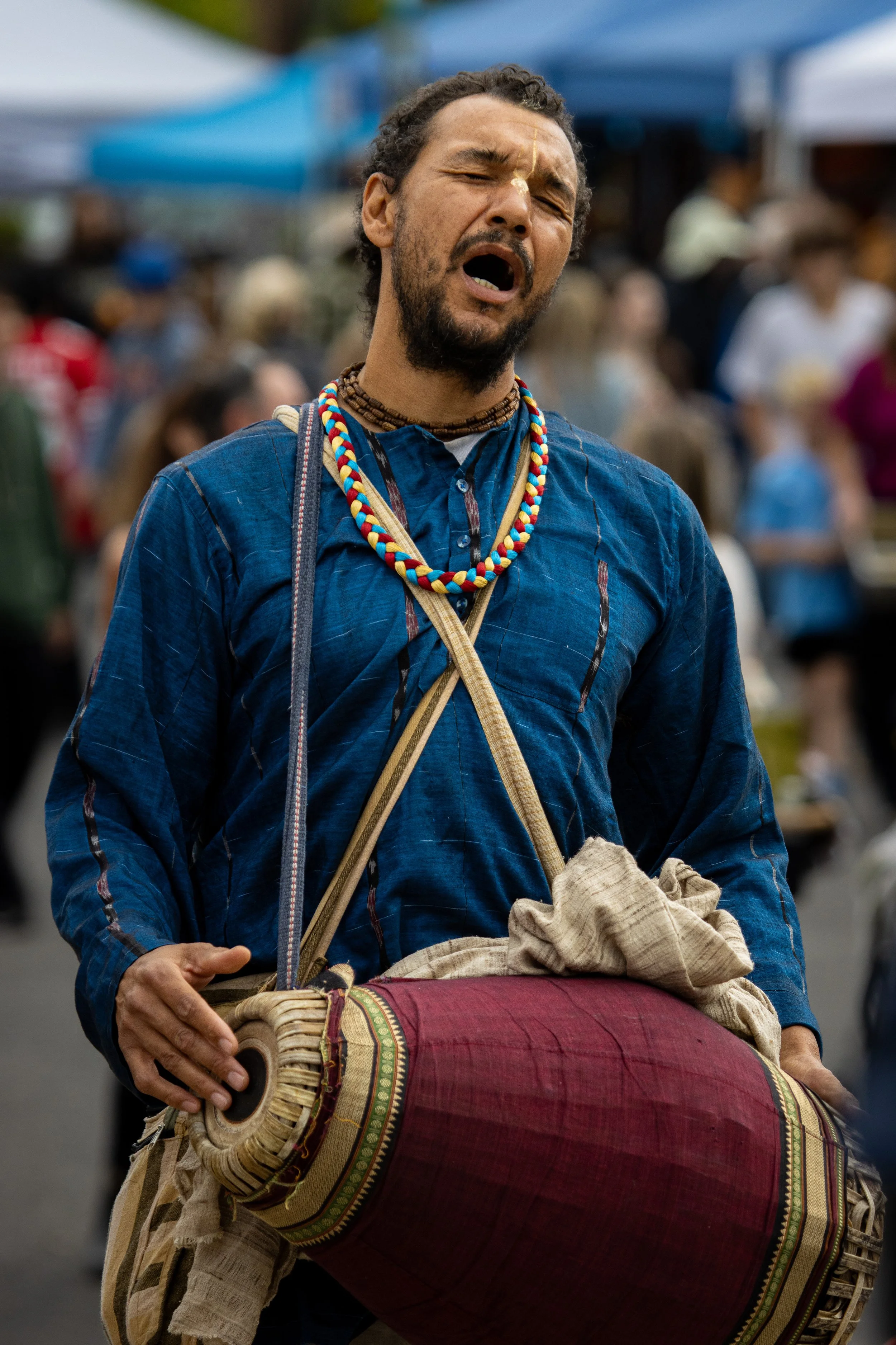 A man with a beard and long hair, wearing a blue shirt and multiple necklaces, appears to be singing or shouting with his eyes shut, at an outdoor event or gathering with a crowd in the background.
