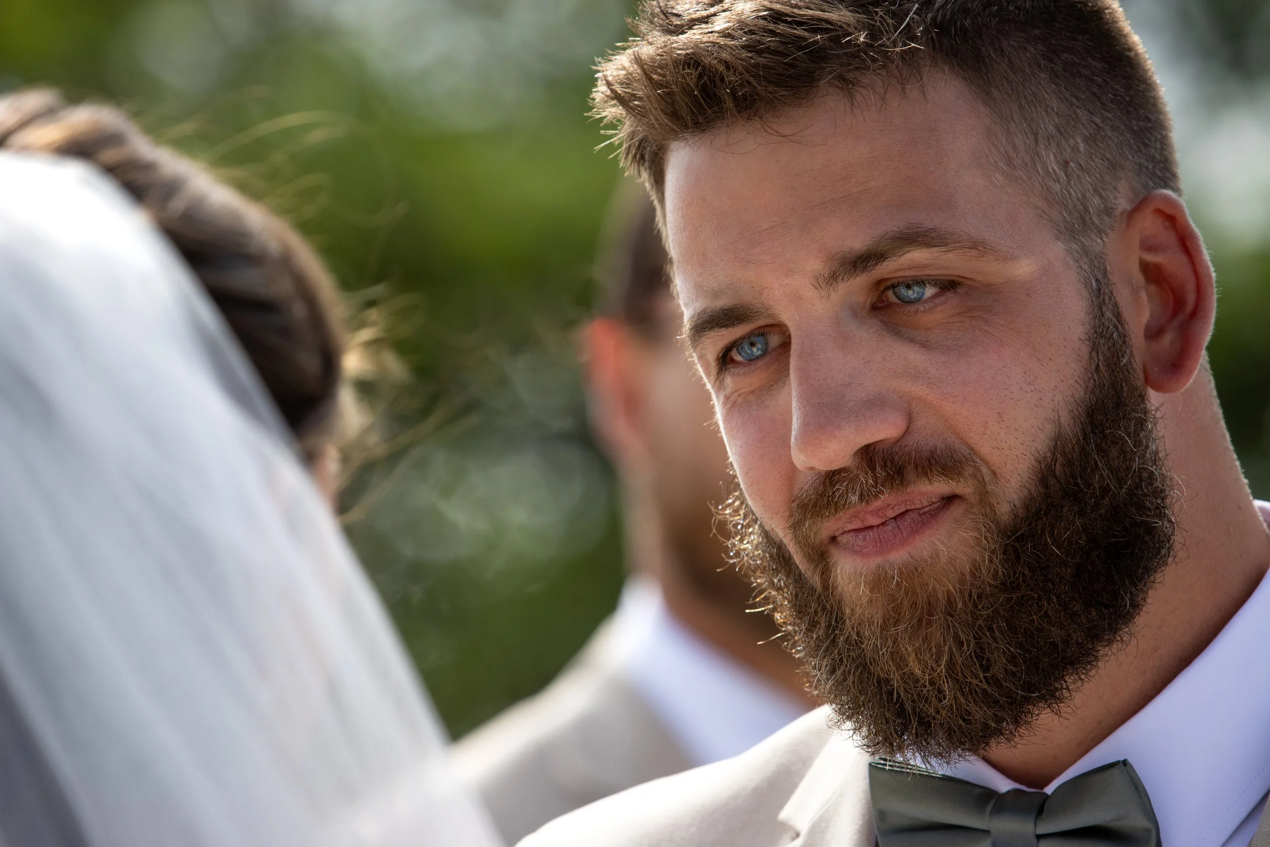 A man with a beard and blue eyes dressed in a formal suit with a bow tie looking at another person.