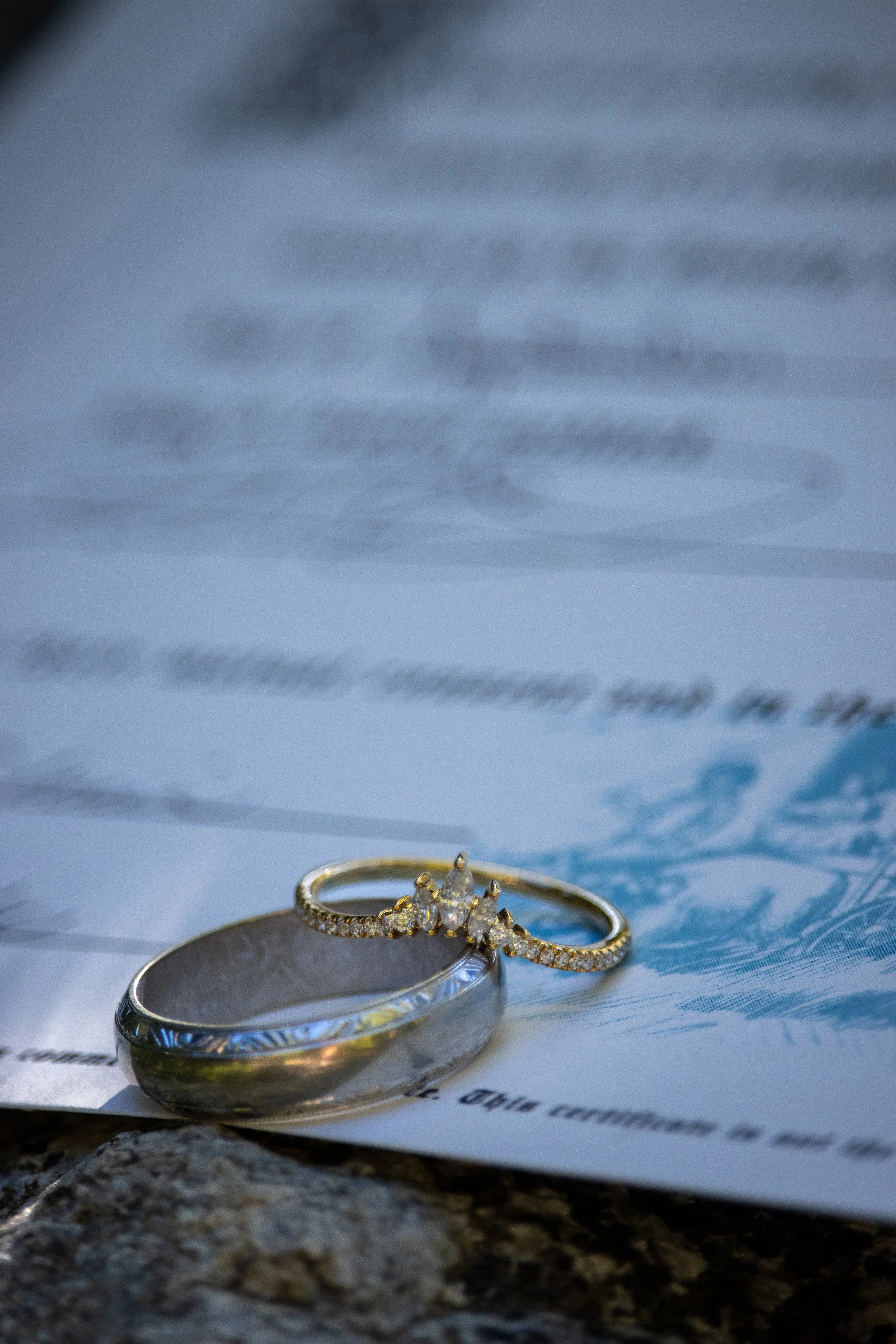 Close-up of two rings, a band and a diamond engagement ring with small diamonds along the band, placed on a piece of paper on a rocky surface. The background is blurred, emphasizing the rings in focus.