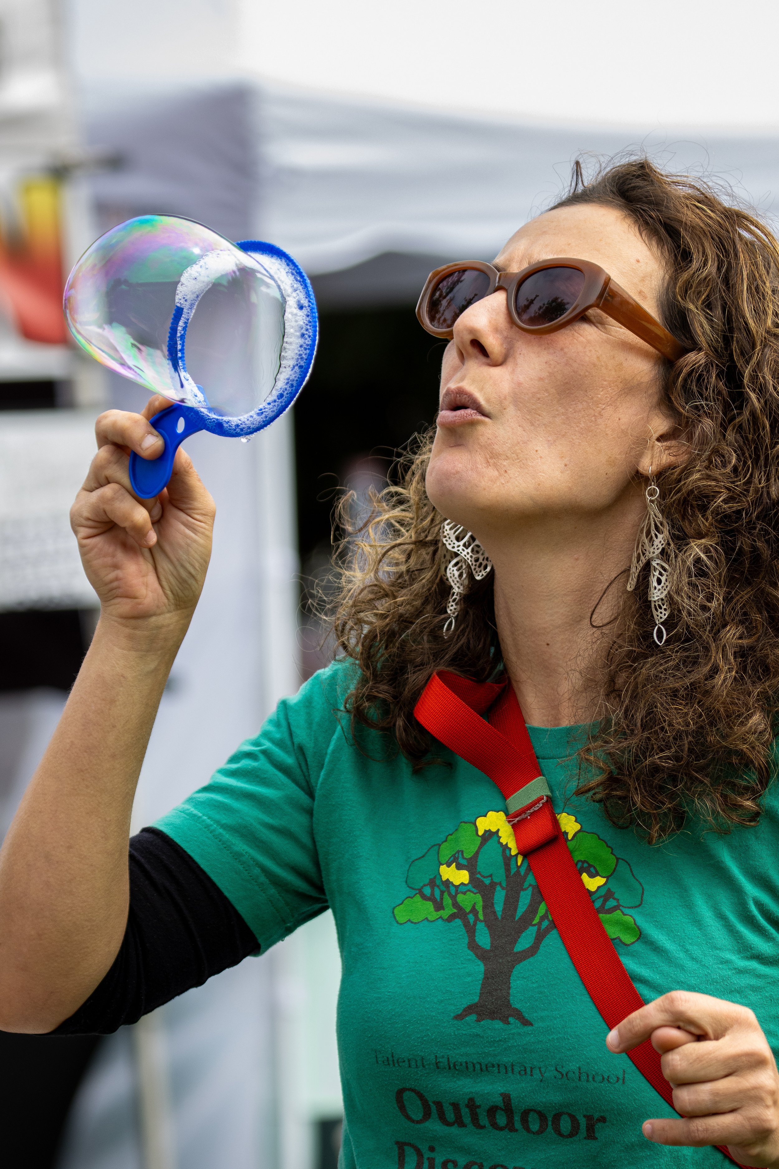 A woman with curly hair wearing sunglasses and a green T-shirt blowing a soap bubble with a blue bubble wand.