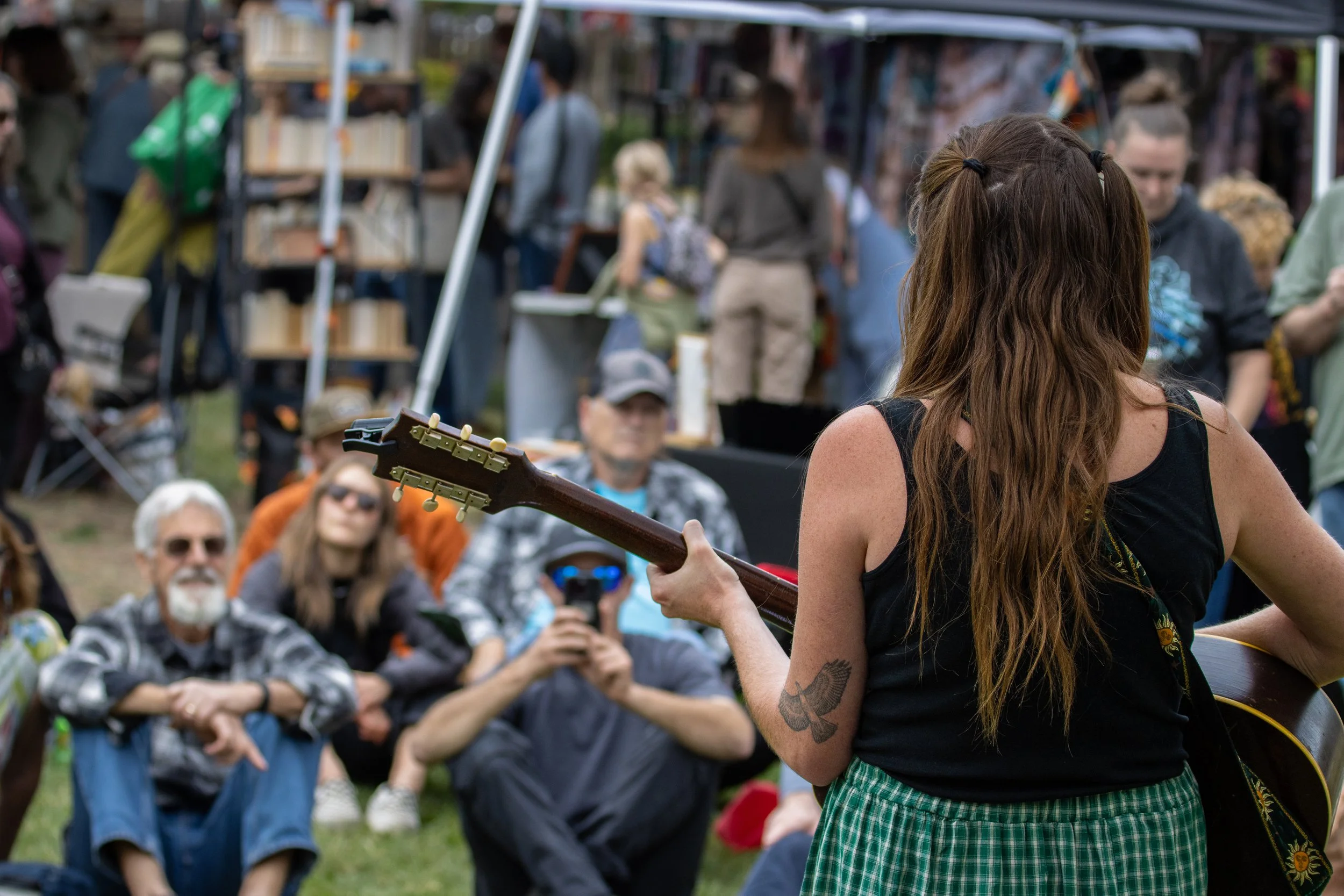 A woman with long brown hair playing an acoustic guitar at an outdoor event, with an audience seated and standing in the background.