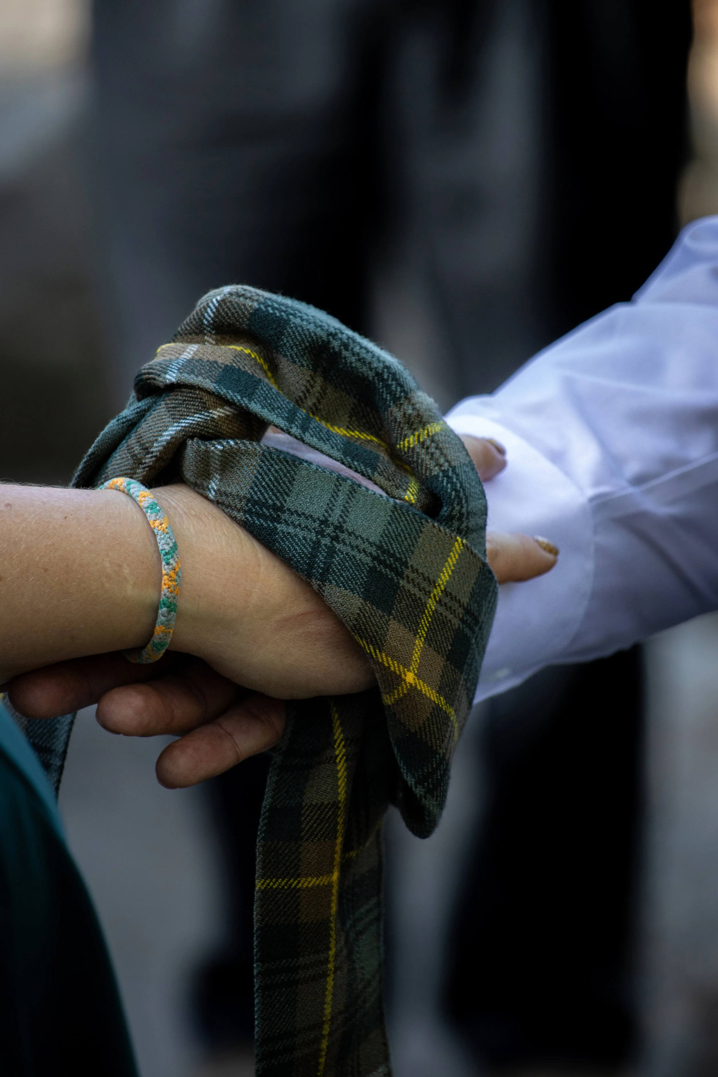 Close-up of two hands holding each other, one hand with a plaid sleeve and the other with a white sleeve, in an outdoor setting.