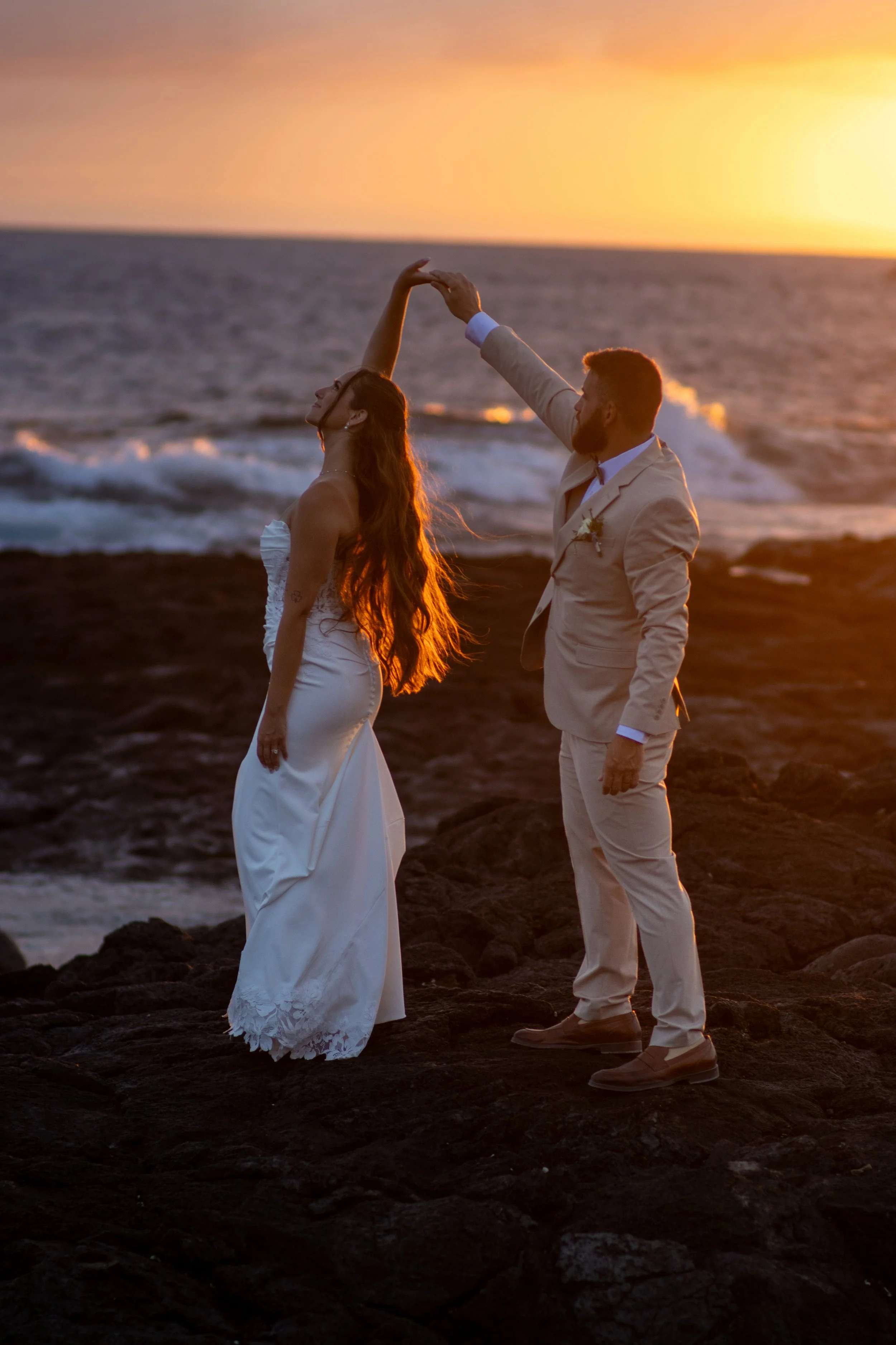 A couple dancing on a rocky beach during sunset, with the woman wearing a white dress and the man in a beige suit.