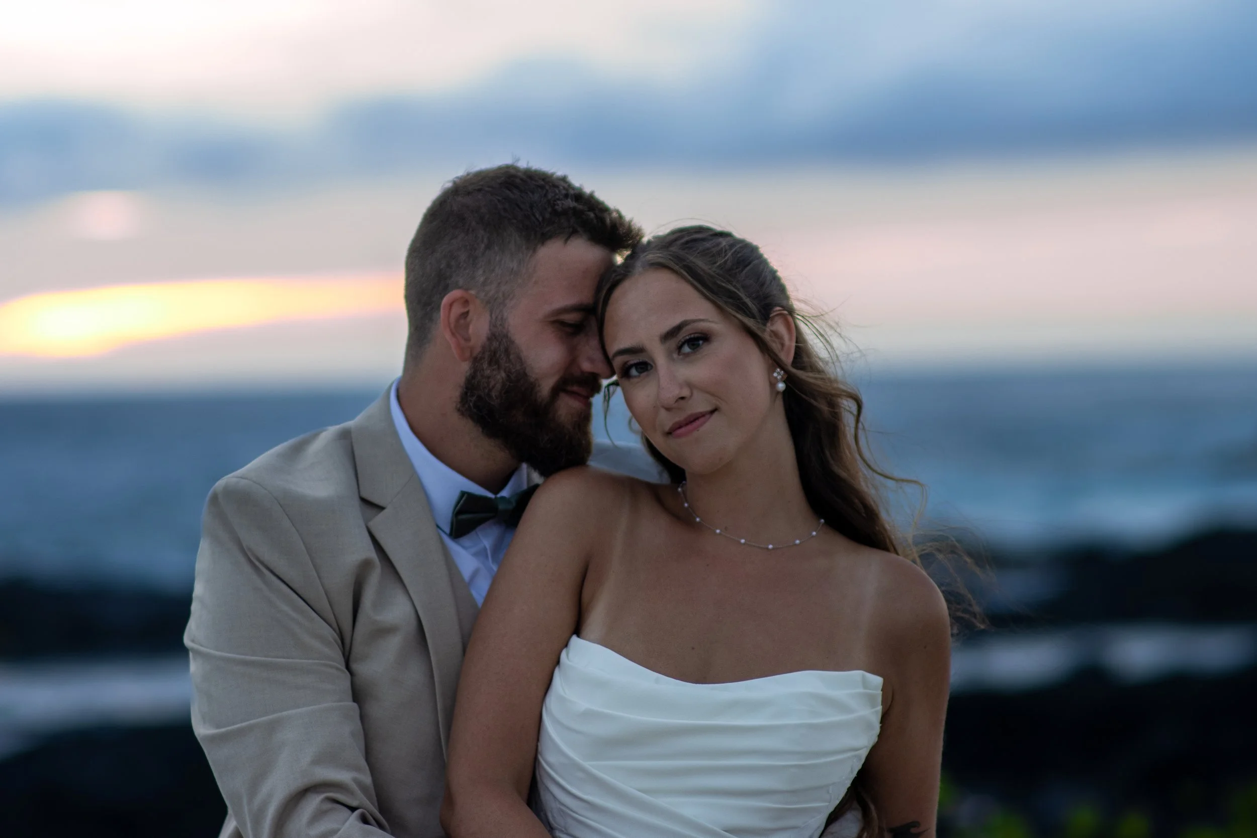 A couple dressed in formal attire, embracing each other outdoors at sunset, with the ocean in the background.