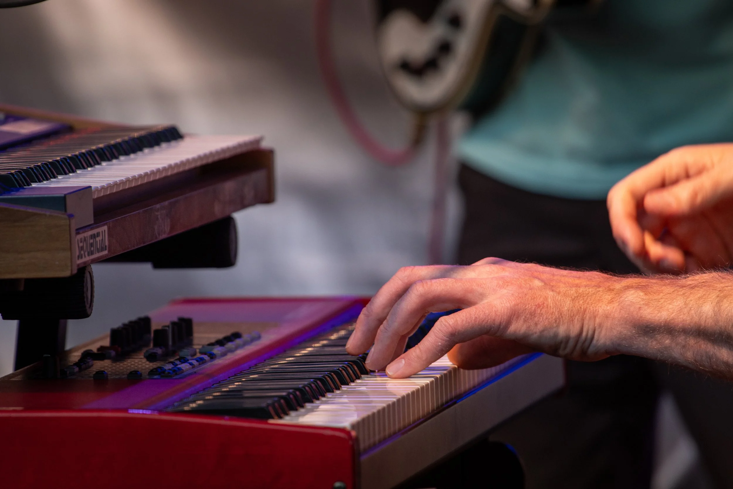 A person playing a red keyboard synthesizer, focusing on their hands on the keys, with a blurred person in the background.