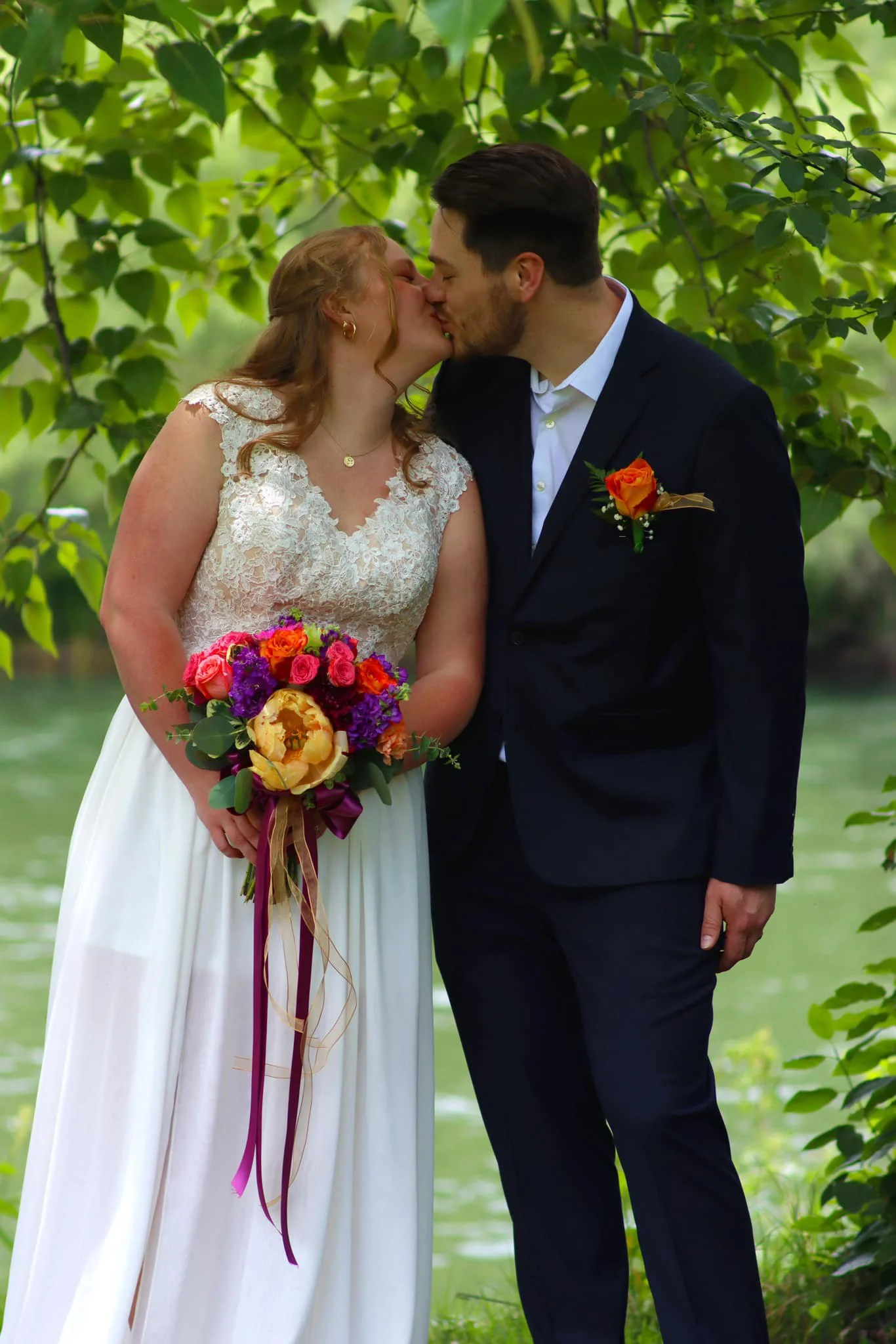 A bride and groom kissing outdoors beneath green tree leaves. The bride holds a colorful bouquet and wears a white lace wedding dress. The groom wears a dark suit with a white shirt and a boutonnière. The setting appears to be near a body of water wi