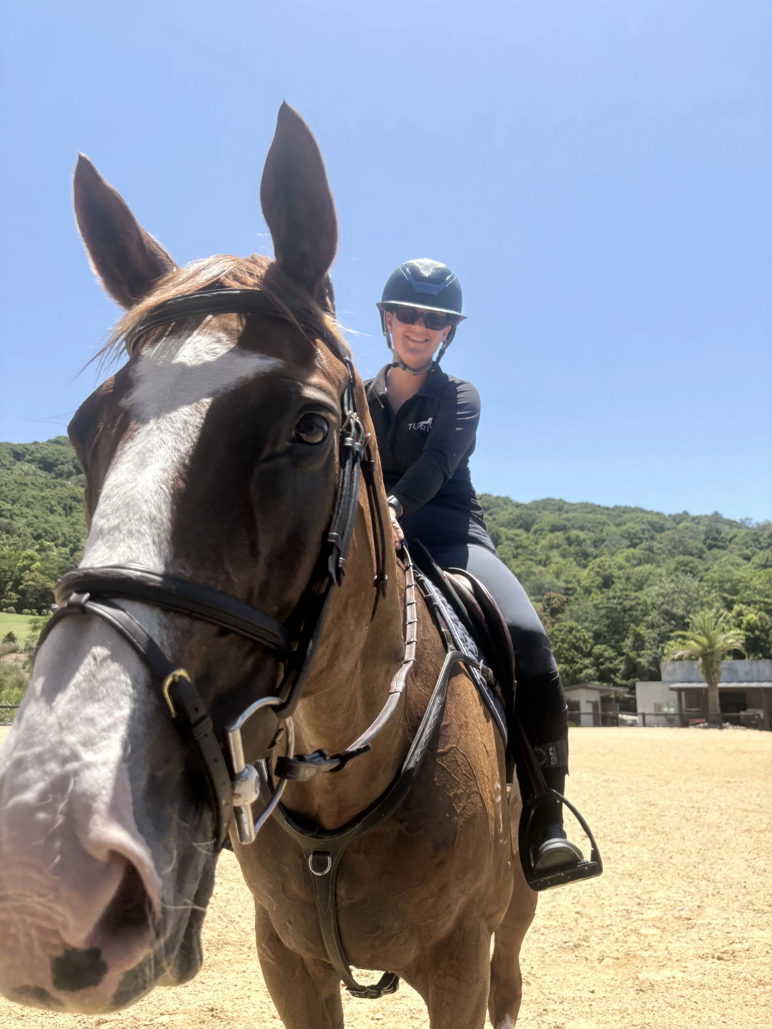 A woman riding a brown horse with white markings on its face, wearing a helmet and sunglasses, on a sunny day in a rural area with green hills and small buildings in the background.
