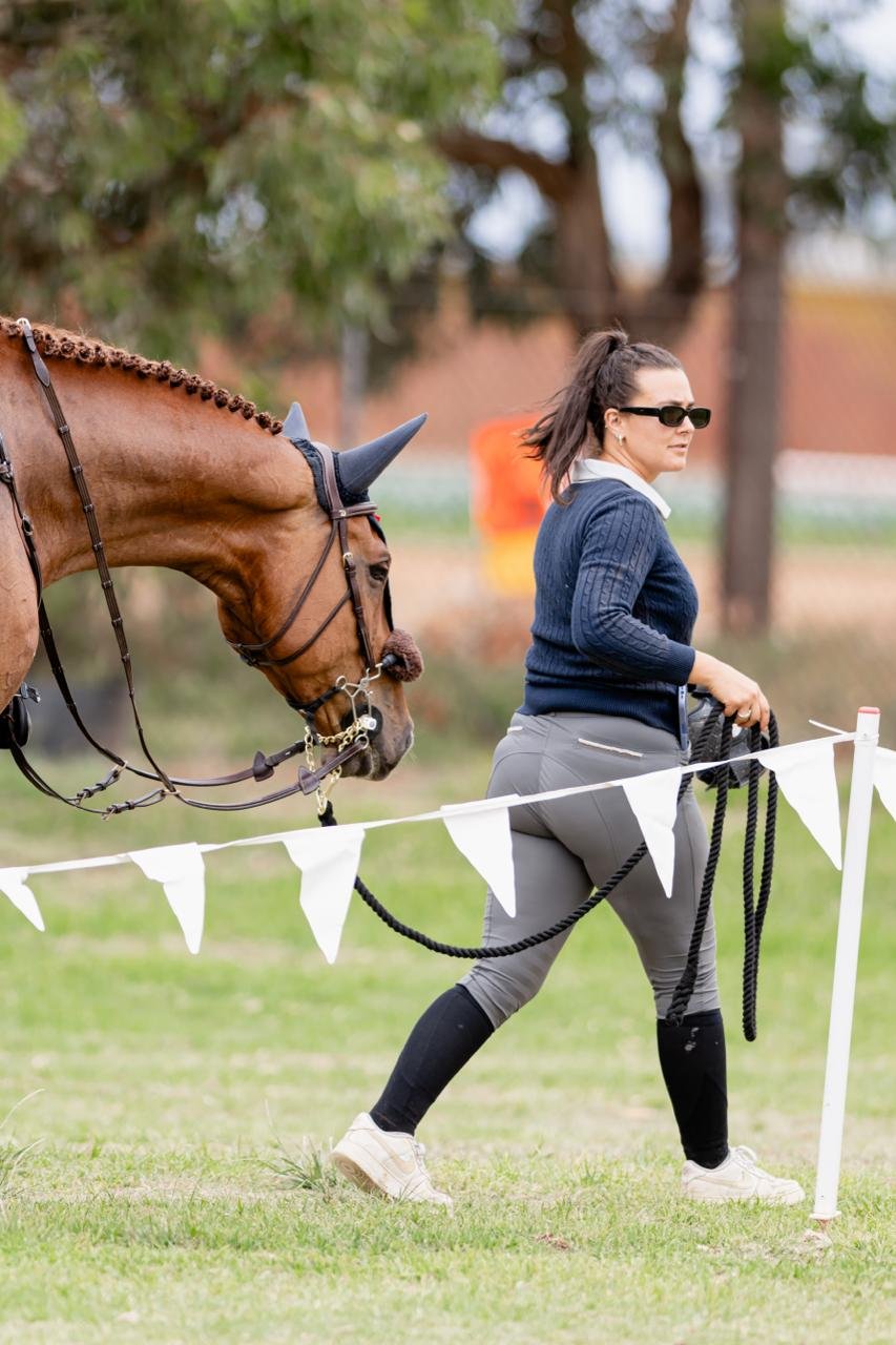 Woman walking a horse in a field, wearing sunglasses, a navy sweater, gray pants, and white shoes.
