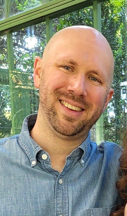 Close-up of a bald man with a beard, smiling, wearing a blue button-up shirt, outdoors near a green tent or greenhouse with trees in the background.