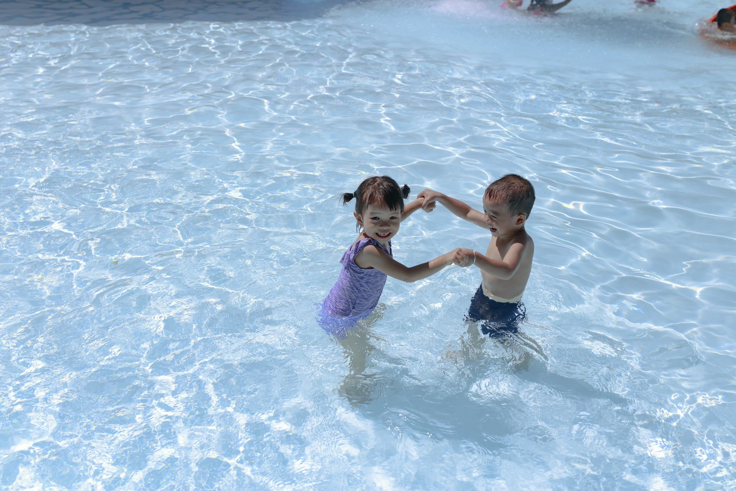 Two children, a girl and a boy, are playing and holding hands in a swimming pool, smiling at each other.