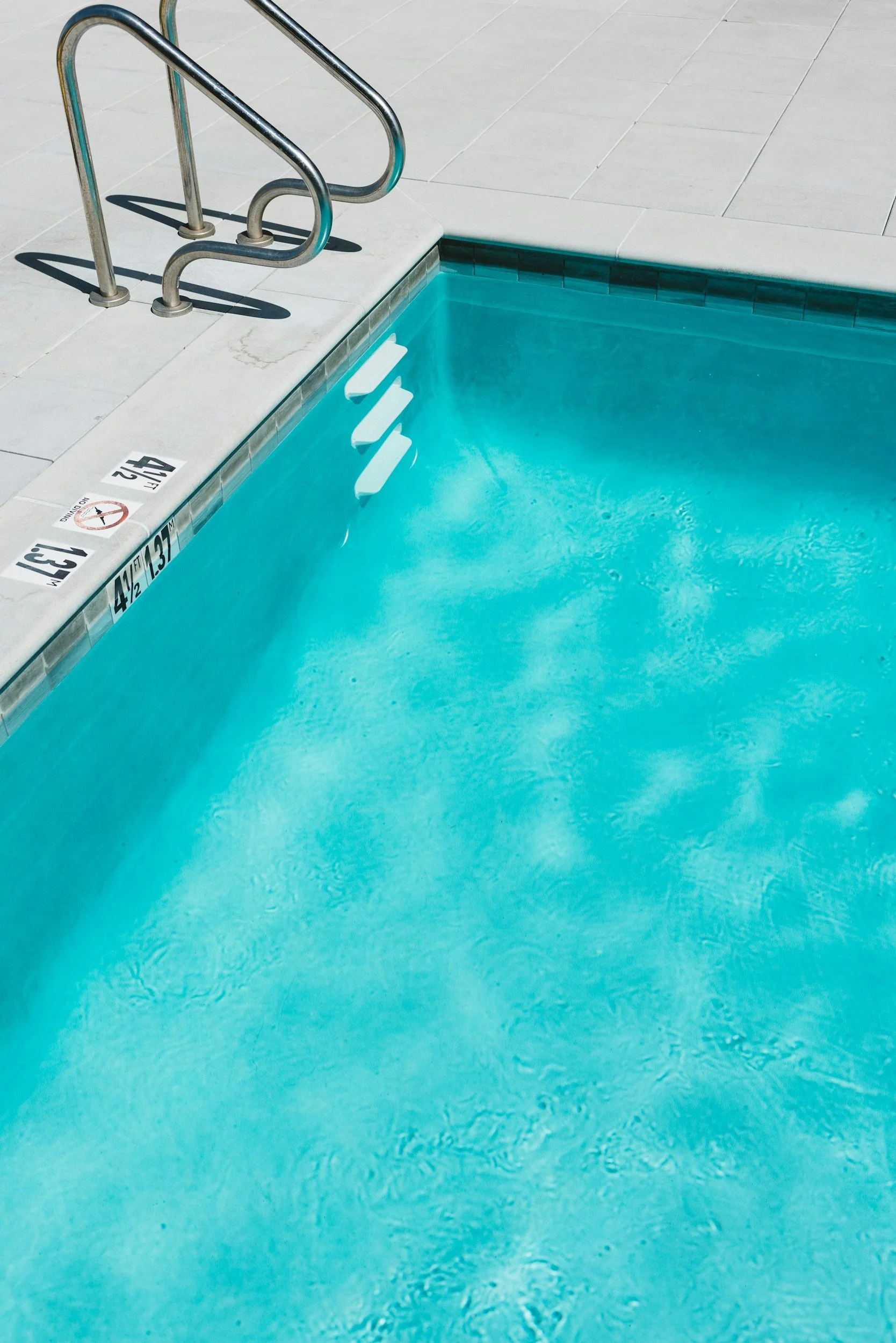 A swimming pool with clear blue water, metallic handrails, and set of three white steps leading into the pool. The pool edge has depth markers and no diving signage visible.