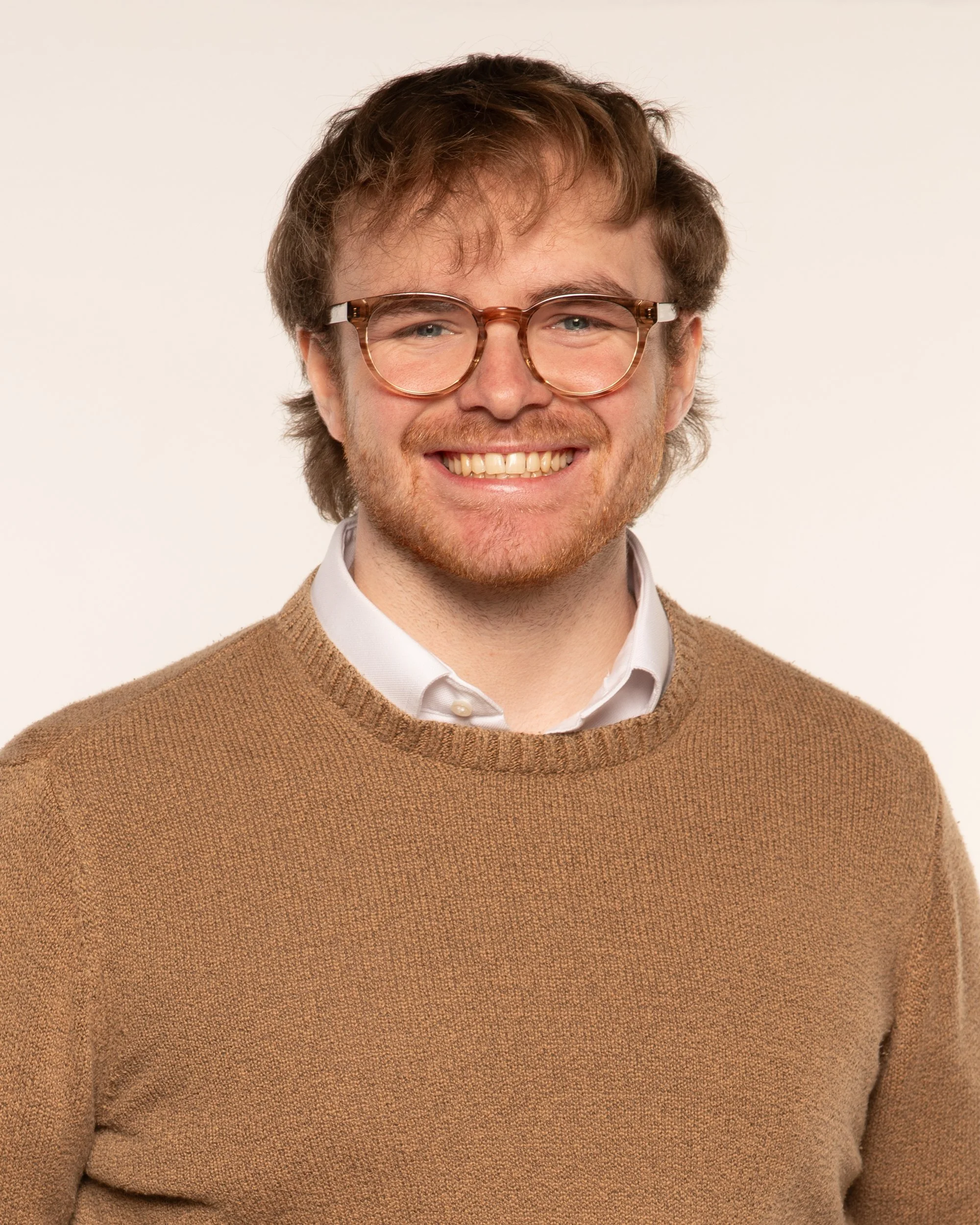 Close-up of a smiling man with brown hair, glasses, wearing a white shirt and brown sweater, against a plain background.
