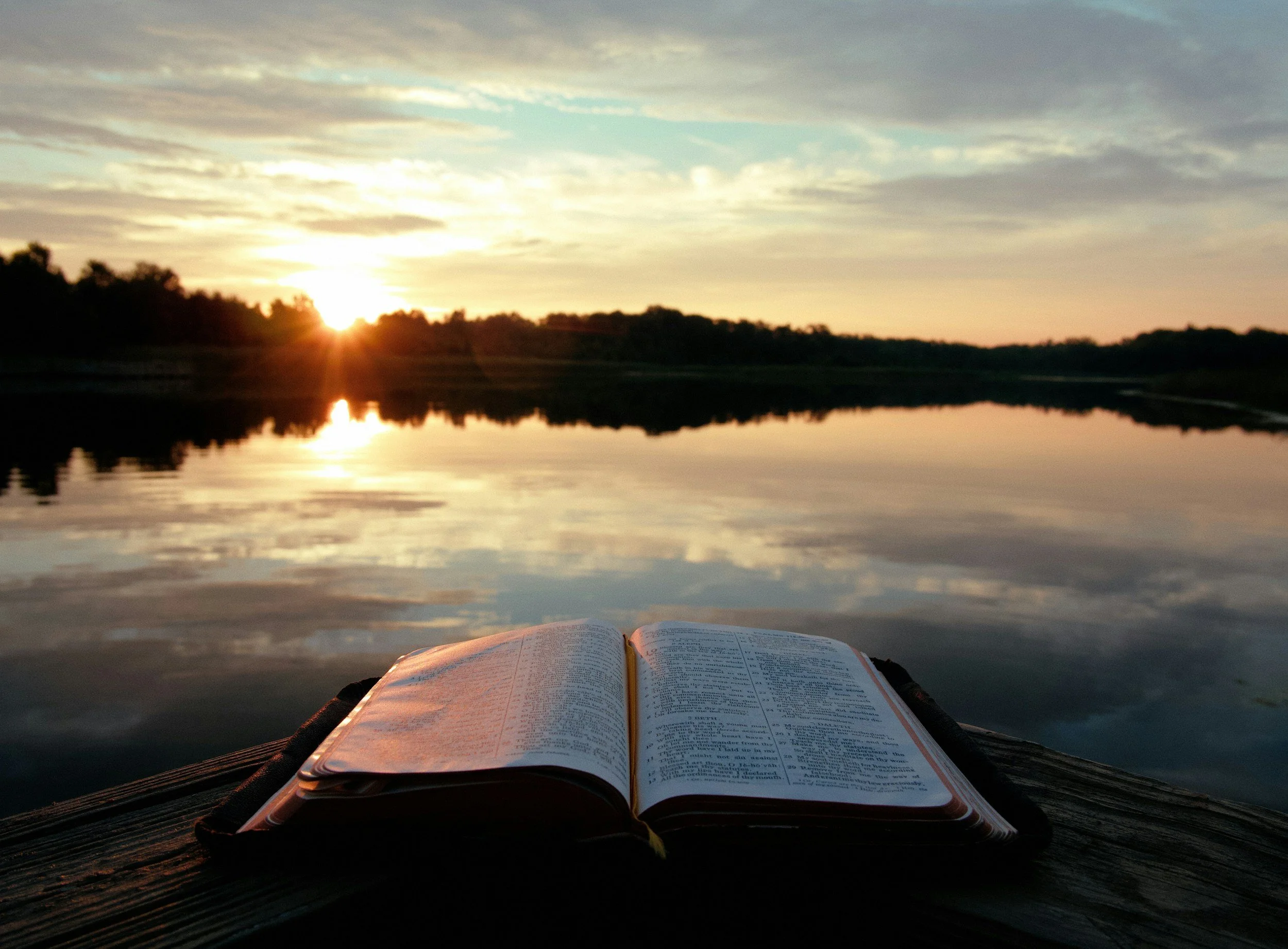 A person is reading the bible during sunrise with a glow from the sun near the horizon, reflected in the water, creating a peaceful and scenic moment.