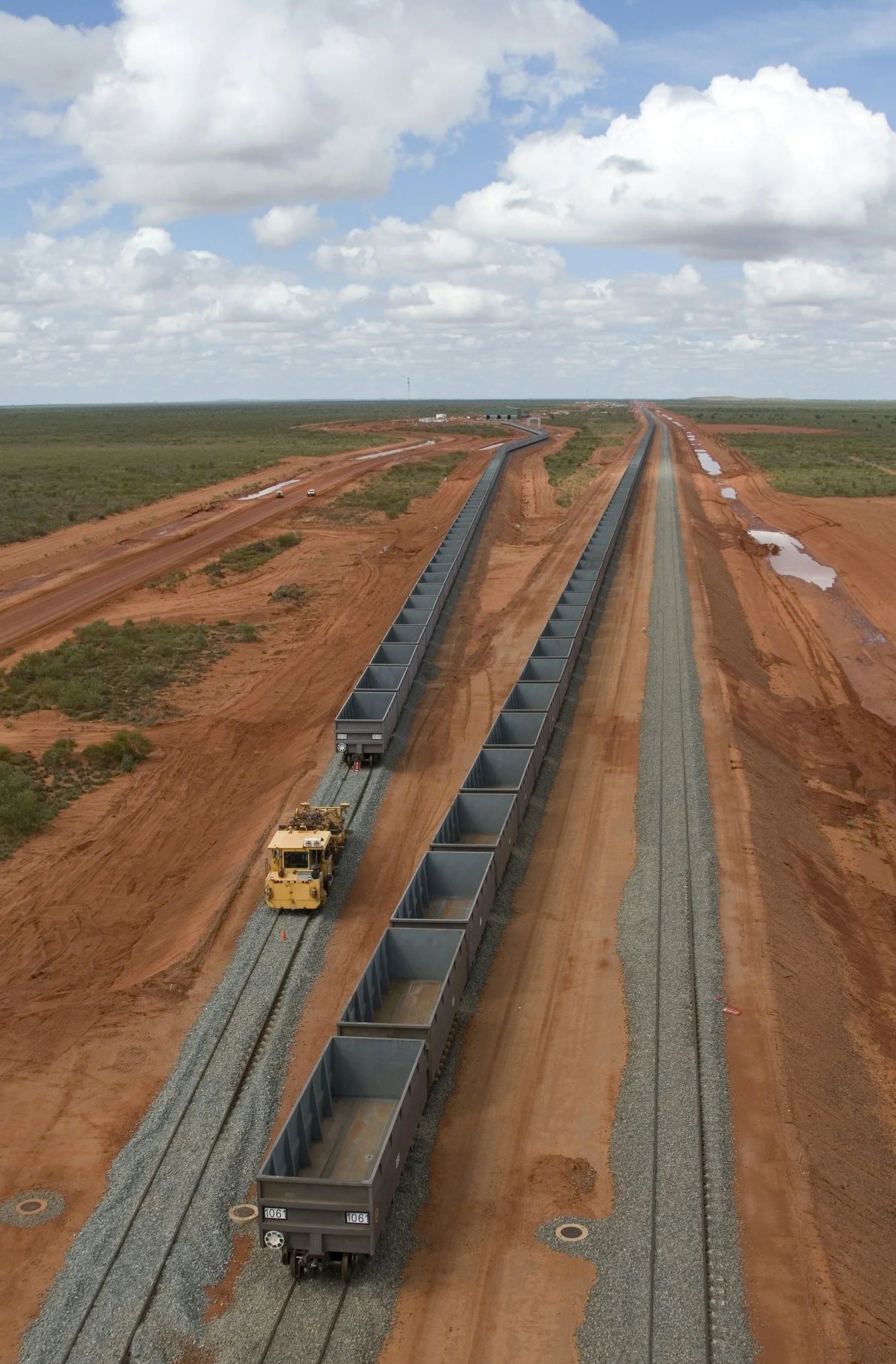 A remote area with red dirt, two tracks, and a train car moving on one of the tracks, with construction machinery nearby and a cloudy sky overhead.