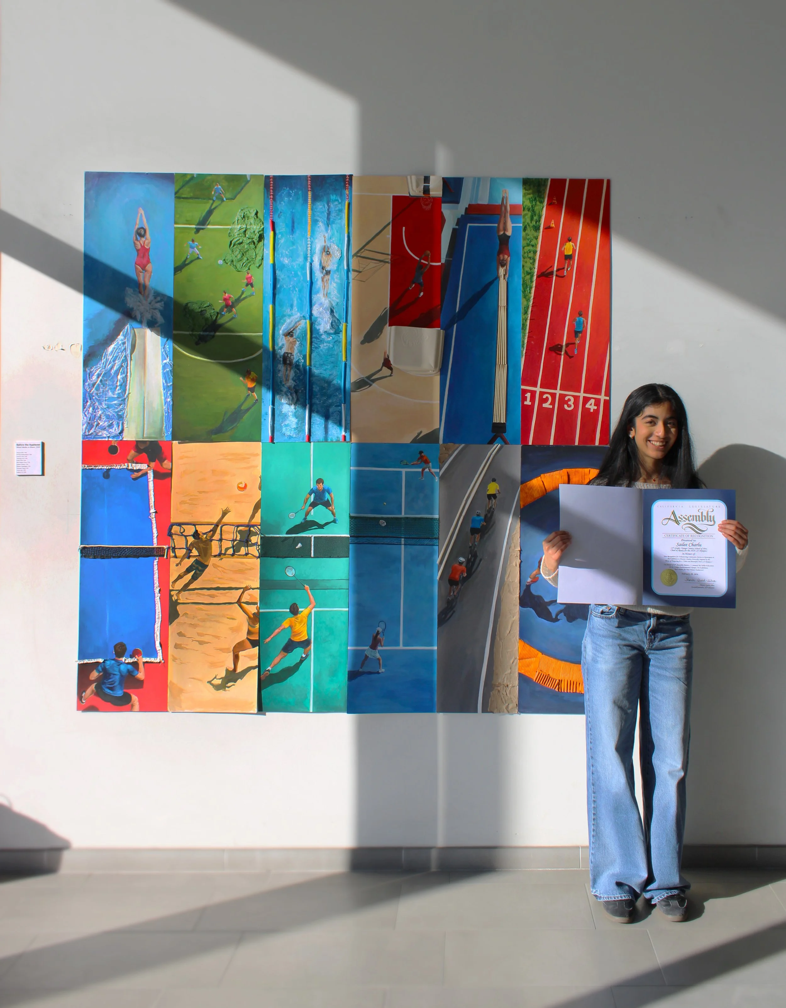 A young woman with long dark hair, wearing jeans and sneakers, standing in front of a wall displaying a collage of sports illustrations, holding a certificate of recognition.