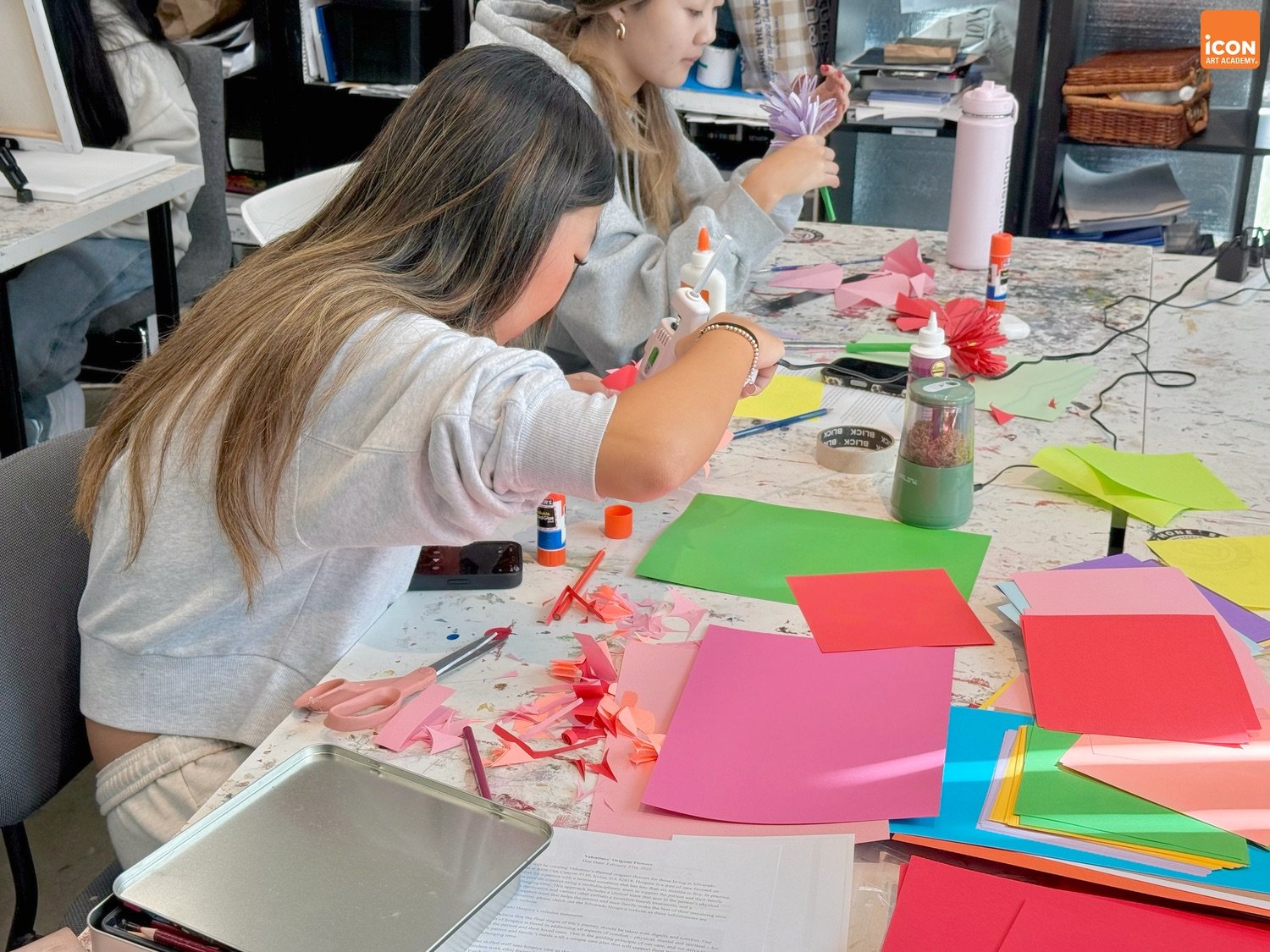 Two young women are sitting at a table engaged in arts and crafts, with colorful paper, glue, scissors, and glue sticks scattered across the table, working on paper flowers or decorations.