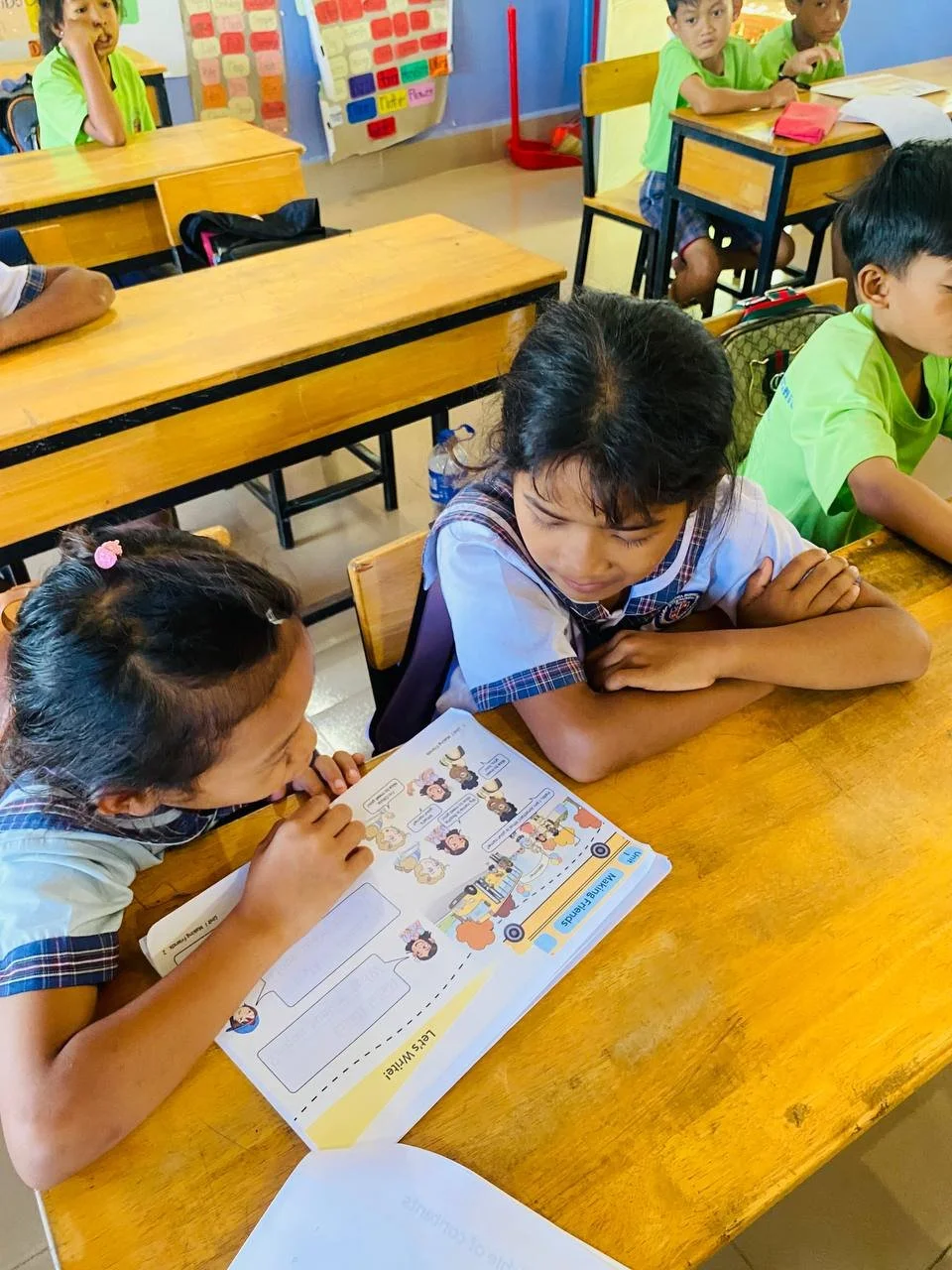 Two young girls in school uniforms reading a workbook at a wooden desk, with other students and classroom decor in the background.