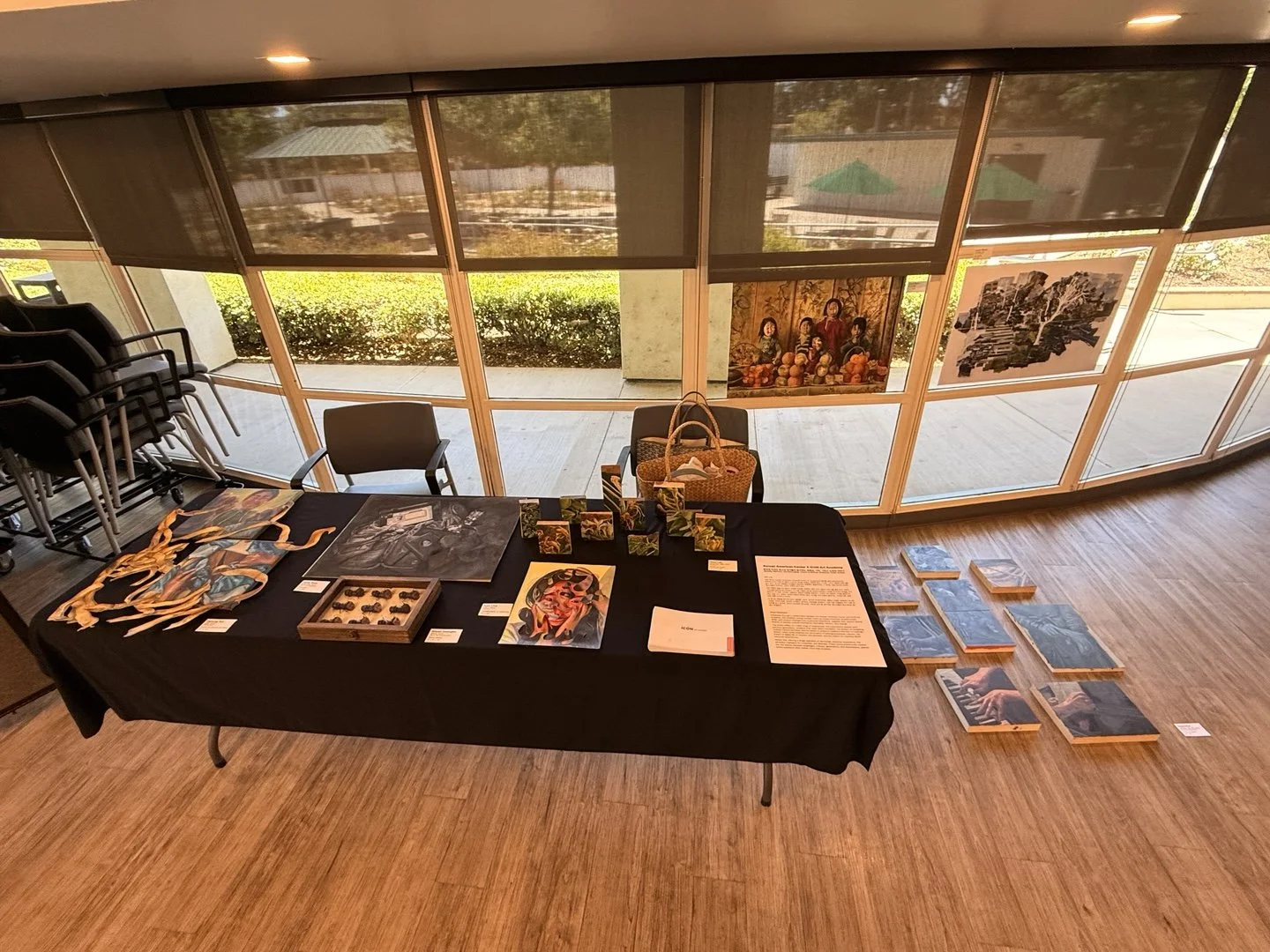 An art exhibit display table with various artwork and items, set in front of large windows with art hanging outside. Artwork includes paintings, small sculptures, and printed images. Chairs, a wicker basket, and stacked chairs are nearby, with additi
