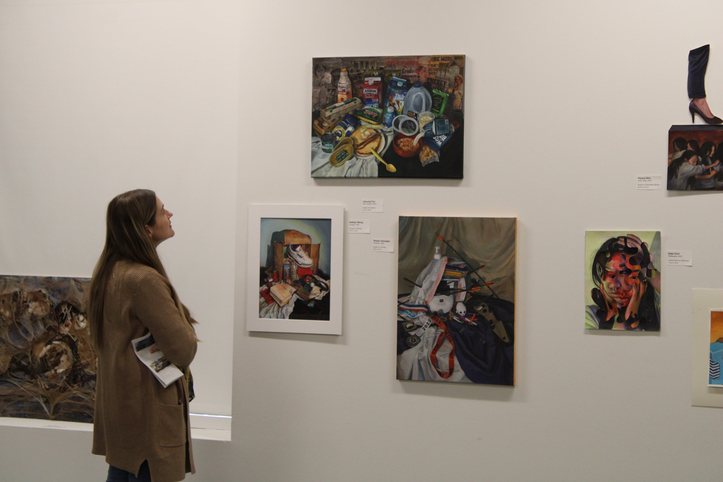 A woman with brown hair in a brown blazer looks at artwork on a museum white wall. The art includes still life paintings and a collage with various objects such as food, a shoe, and toys.