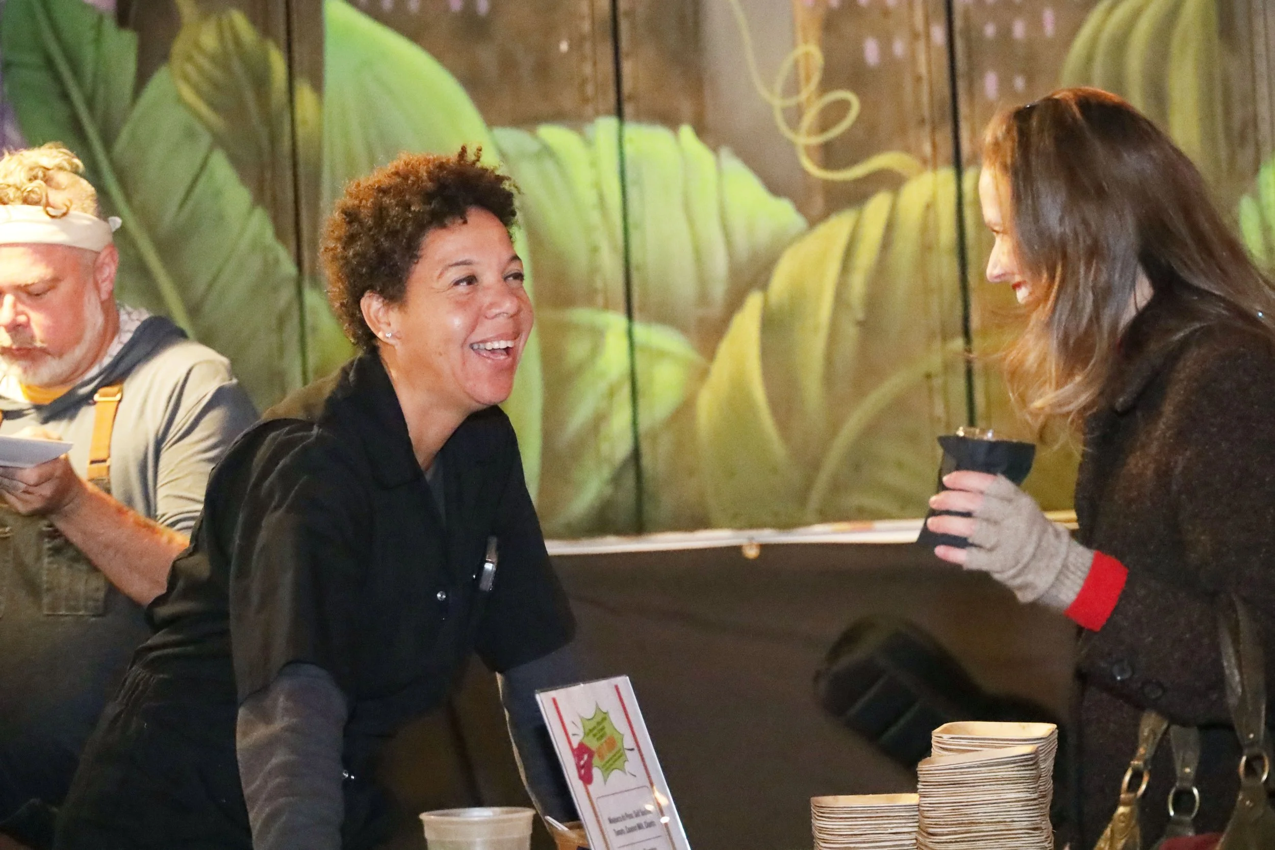 Two women smiling and talking at a food event vendor table.