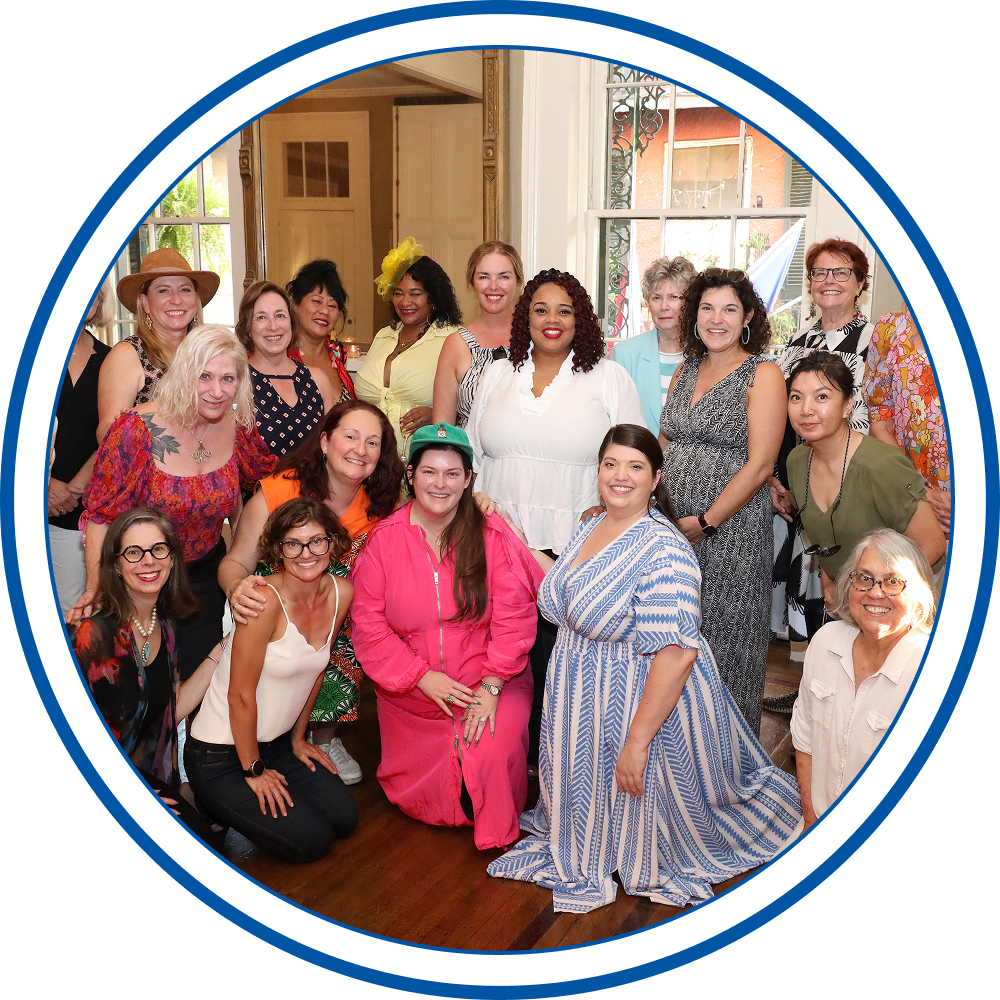 Group of women smiling indoors at a celebration, some wearing colorful dresses and hats, in front of large windows with sunlight.