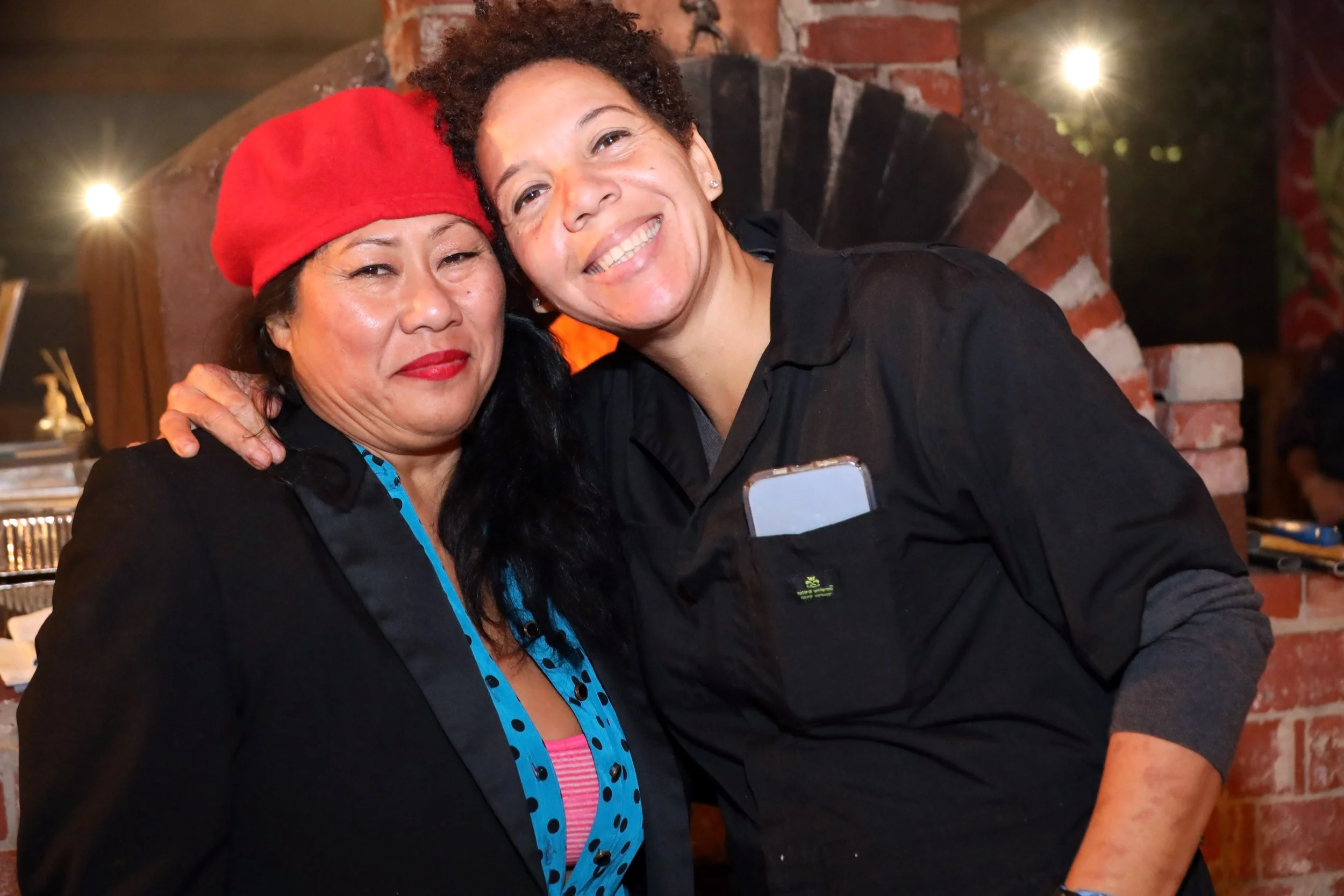 Two women smiling and hugging at an evening event with a brick fireplace in the background. One woman is wearing a red hat and a black jacket, while the other has curly hair and is wearing a black jacket.