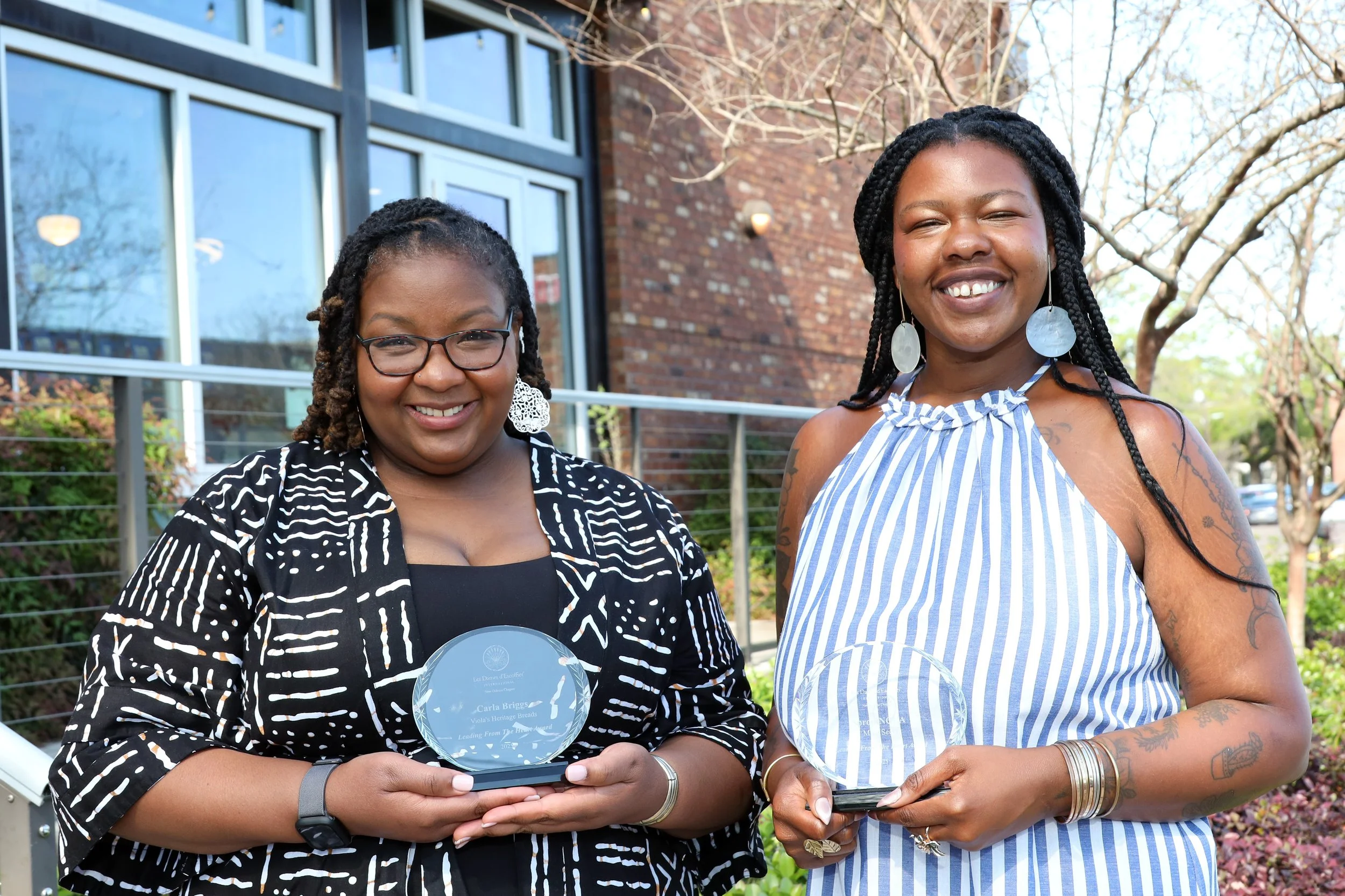 Two women standing outdoors, holding Leading From the Heart Awards, in front of a brick building with large windows. One woman is wearing a black and white patterned top and the other woman is smiling, wearing a blue and white striped dress.
