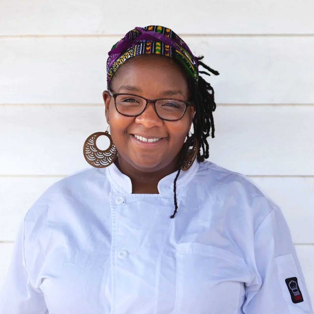 A woman wearing glasses, large earrings, a colorful headwrap, and a white chef's coat, smiling in front of a white wooden wall.