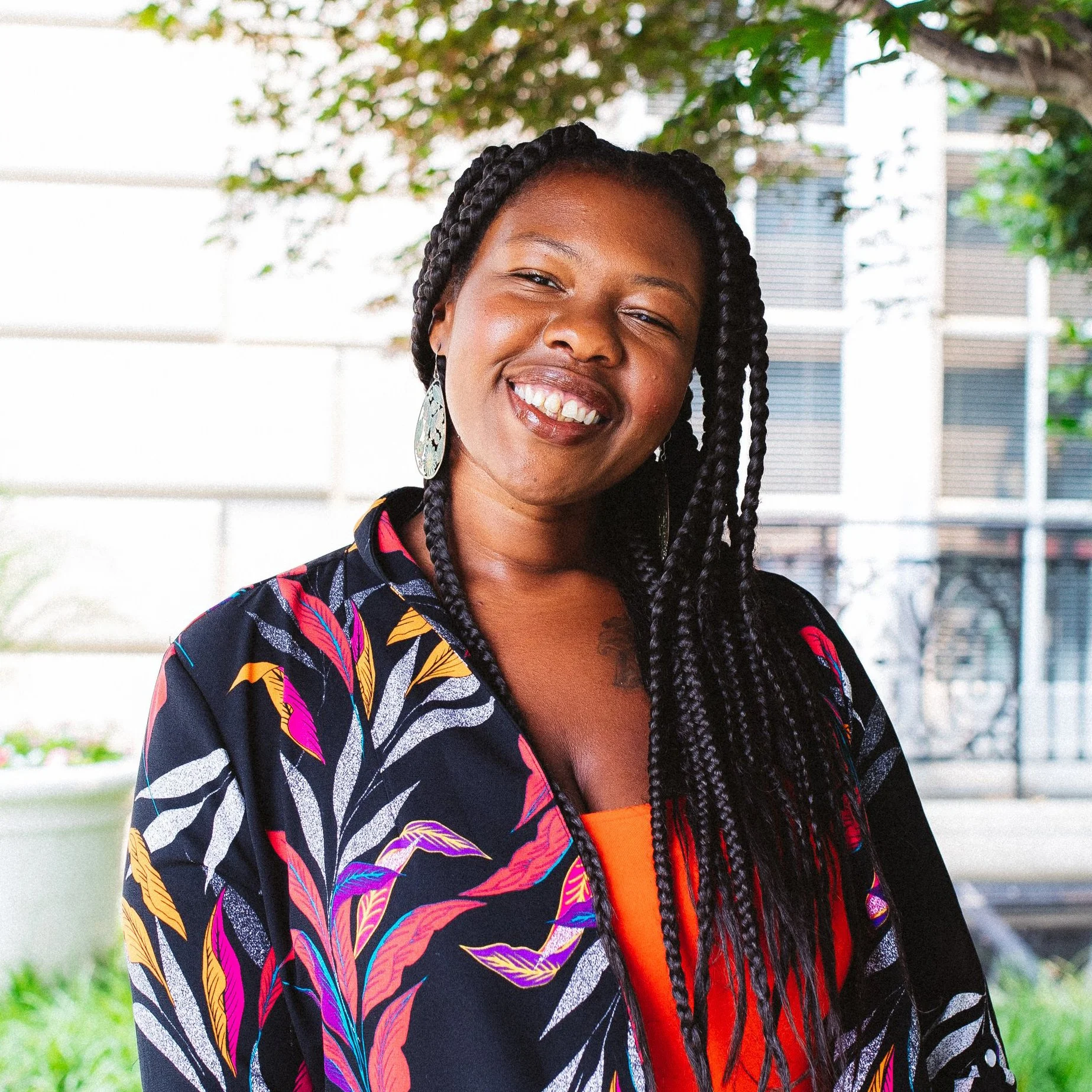 A young woman smiling outdoors with a background of a white fence, trees, and a modern building. She has long braided hair, large earrings, and is wearing a colorful, patterned jacket.