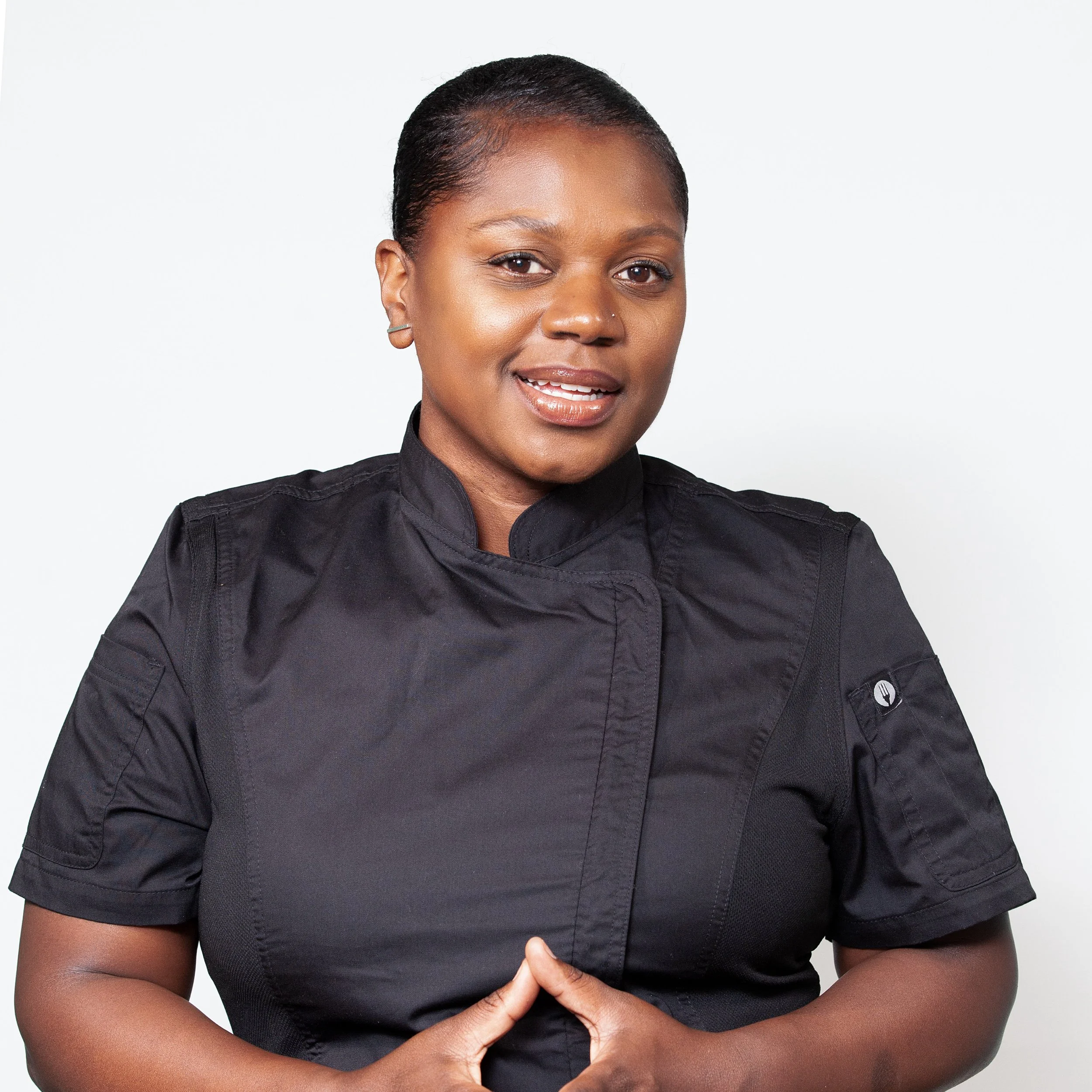 Close-up of a woman wearing a black chef uniform with a small emblem of a fork and spoon on the pocket, smiling, with her hands clasped in front of her.