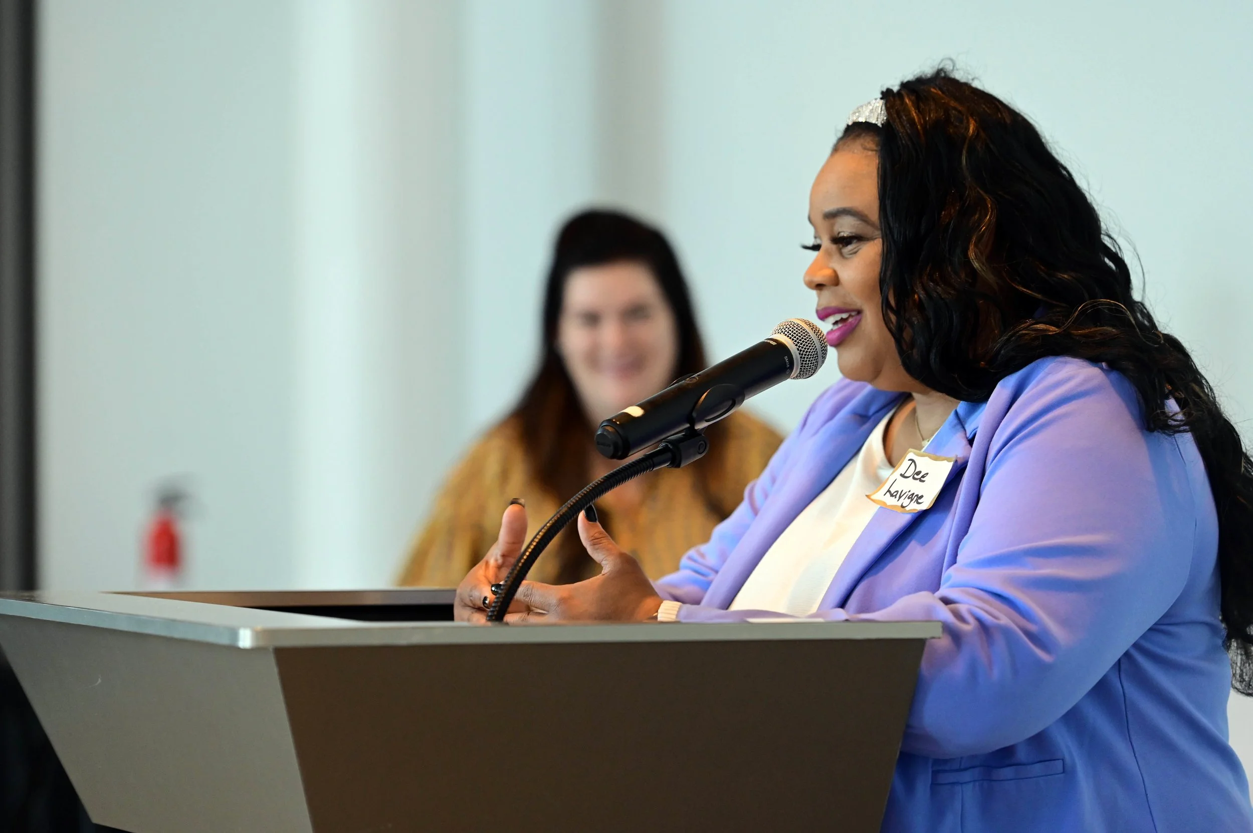 A woman with dark, wavy hair speaking into a microphone at an awards breakfast, wearing a light purple blazer. A woman with reddish-brown hair in the background, smiling.