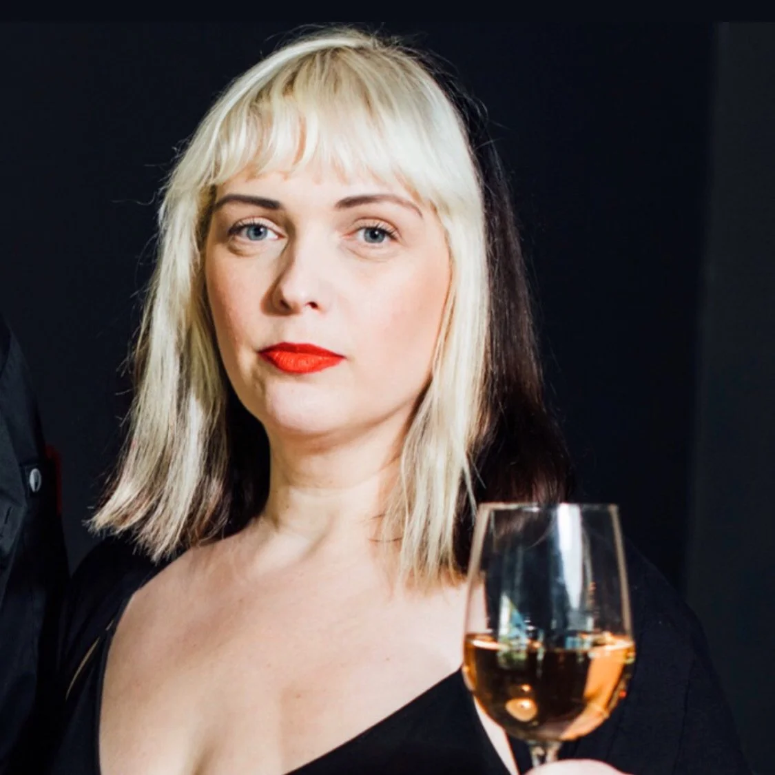 A woman with blonde and dark hair, wearing red lipstick, holding a glass of rosé wine, against a dark background.