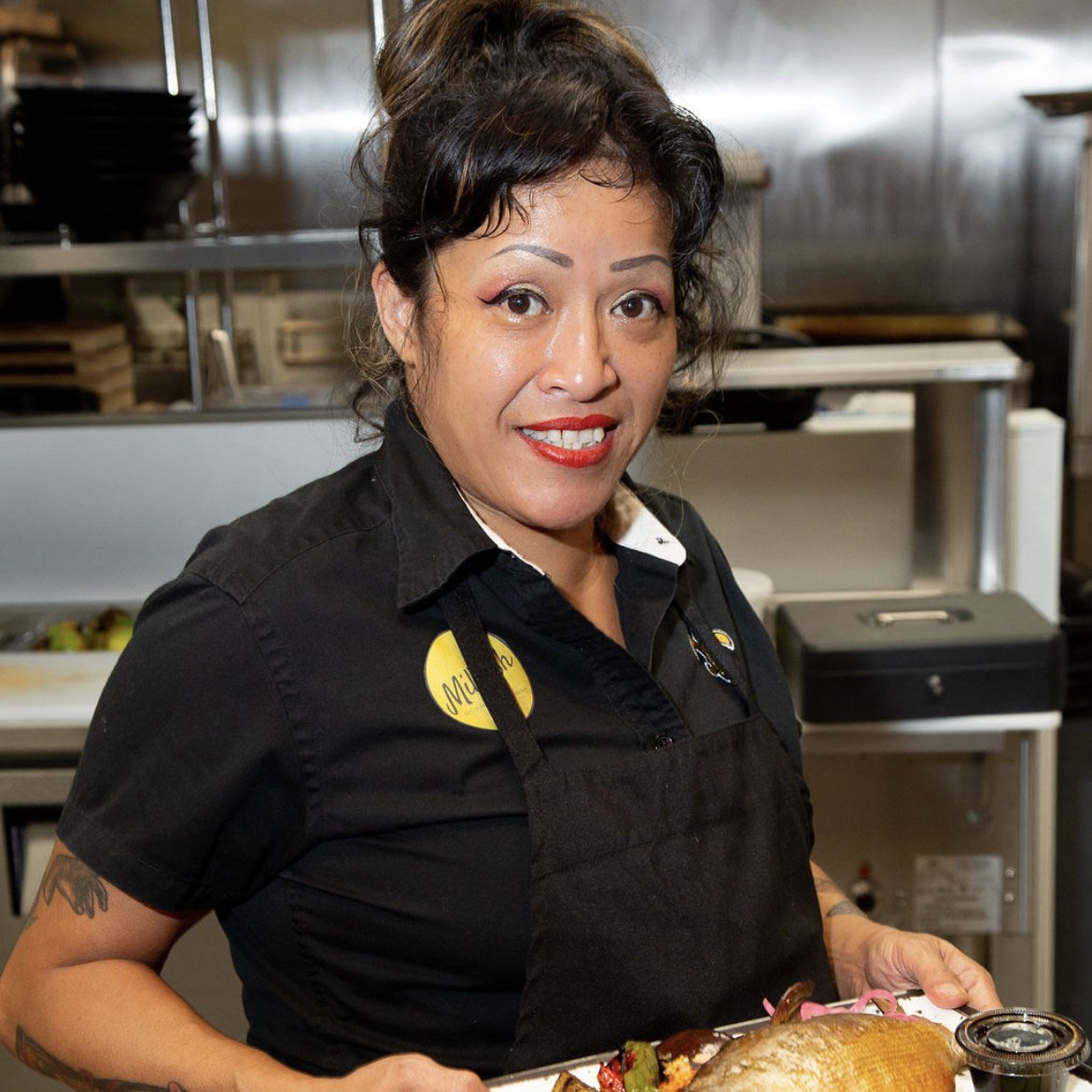 A woman with dark, curly hair, wearing a black shirt, red lipstick, and a black apron, smiling and holding a tray with food in a commercial kitchen.