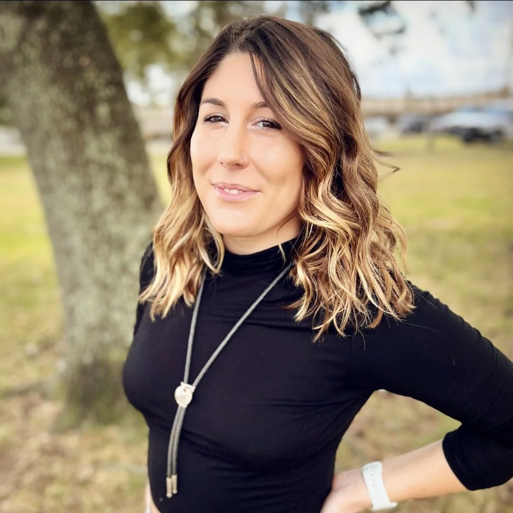A woman with shoulder-length wavy hair, wearing a black top and a silver necklace, standing outdoors next to a tree with her hand on her hip.