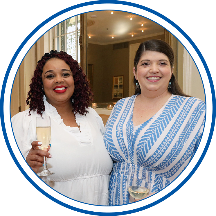 Two women smiling at a social gathering, holding glasses of white wine, standing indoors in front of a mirror and candlelit background.