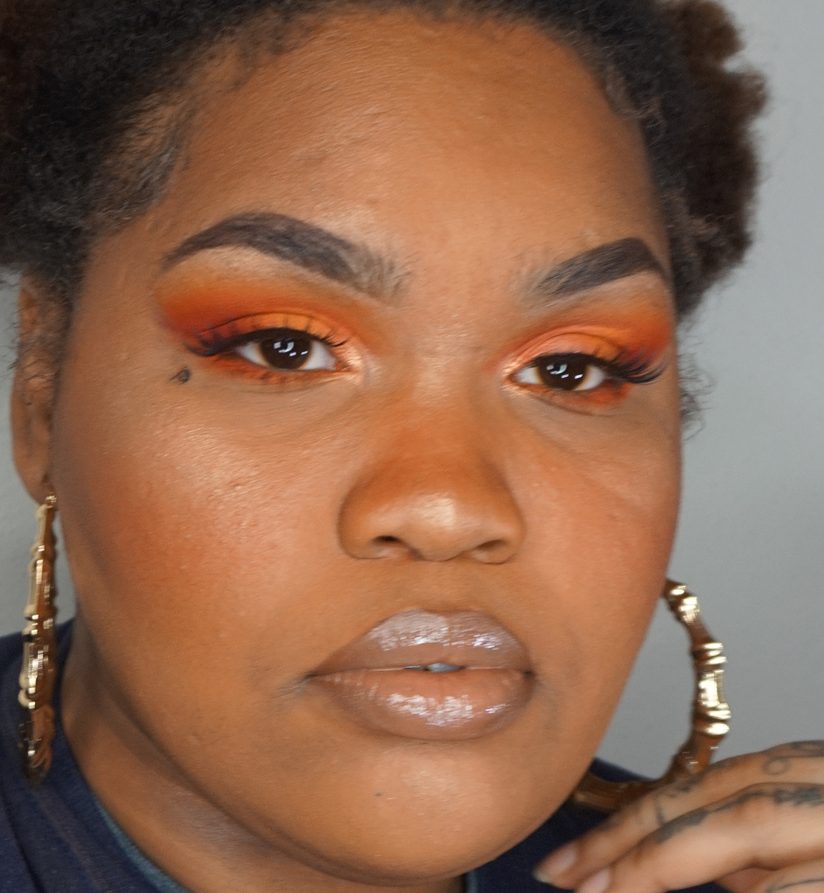 A close-up of a woman with beautifully styled makeup featuring orange and gold eyeshadows, thick eyelashes, and glossy lips, wearing large hoop earrings.