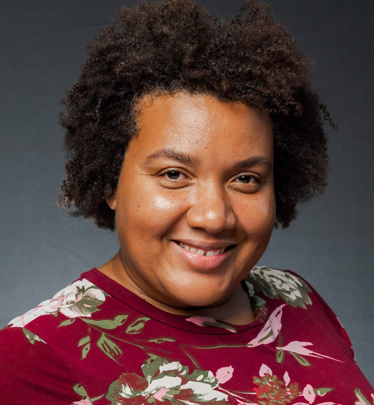 A woman with dark, curly hair smiling, wearing a red floral top against a dark gray background.