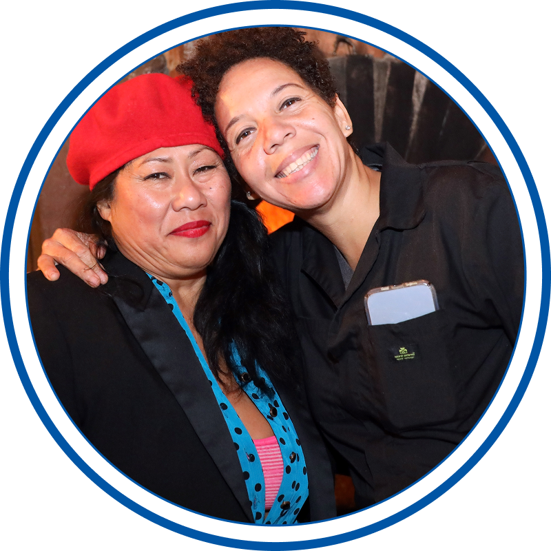 Two women smiling and hugging each other, one wearing a red hat and the other with short curly hair, dressed in black, in an evening outdoor setting.