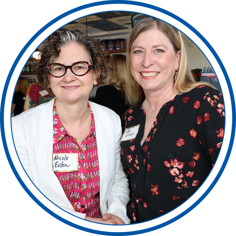Two women smiling, wearing name tags, at a social event. One has curly hair, glasses, and a white blazer over a pink floral shirt. The other has straight blonde hair, earrings, and a black floral dress.